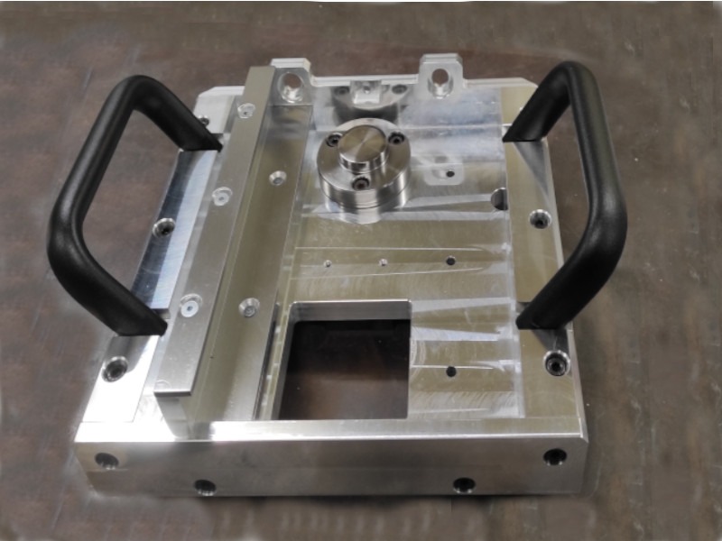 Precyzyjnie obrobiony, aluminiowy element z uchwytami z tworzywa sztucznego, widoczne ślady obróbki i otwory montażowe.