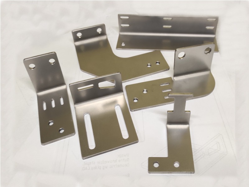 Zestaw precyzyjnie wykonanych, metalowych wsporników i łączników o różnych kształtach, z otworami i szczelinami, ułożonych na białym tle z widocznym fragmentem rysunku technicznego.