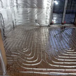 Białe rury ogrzewania podłogowego ułożone na folii z siatką, ściany wyłożone folią termoizolacyjną, widoczne okno.