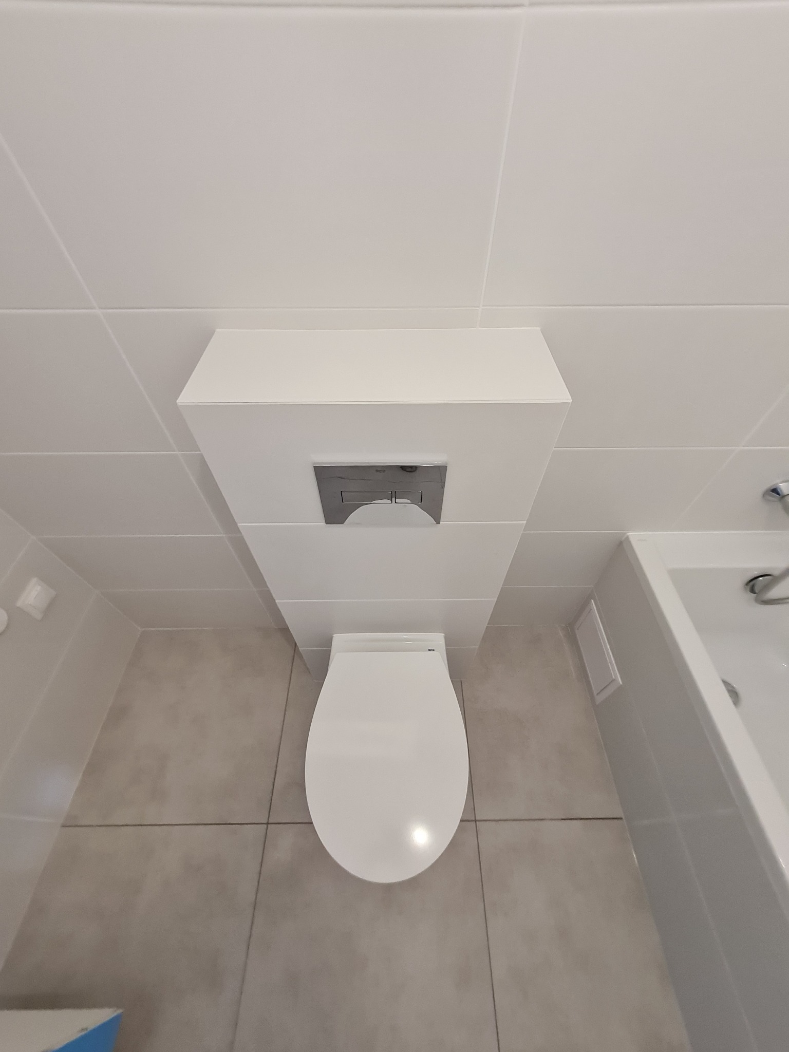 Nowoczesna łazienka z podwieszaną toaletą i zabudowaną spłuczką, widok z góry, białe płytki ścienne i szare podłogowe.