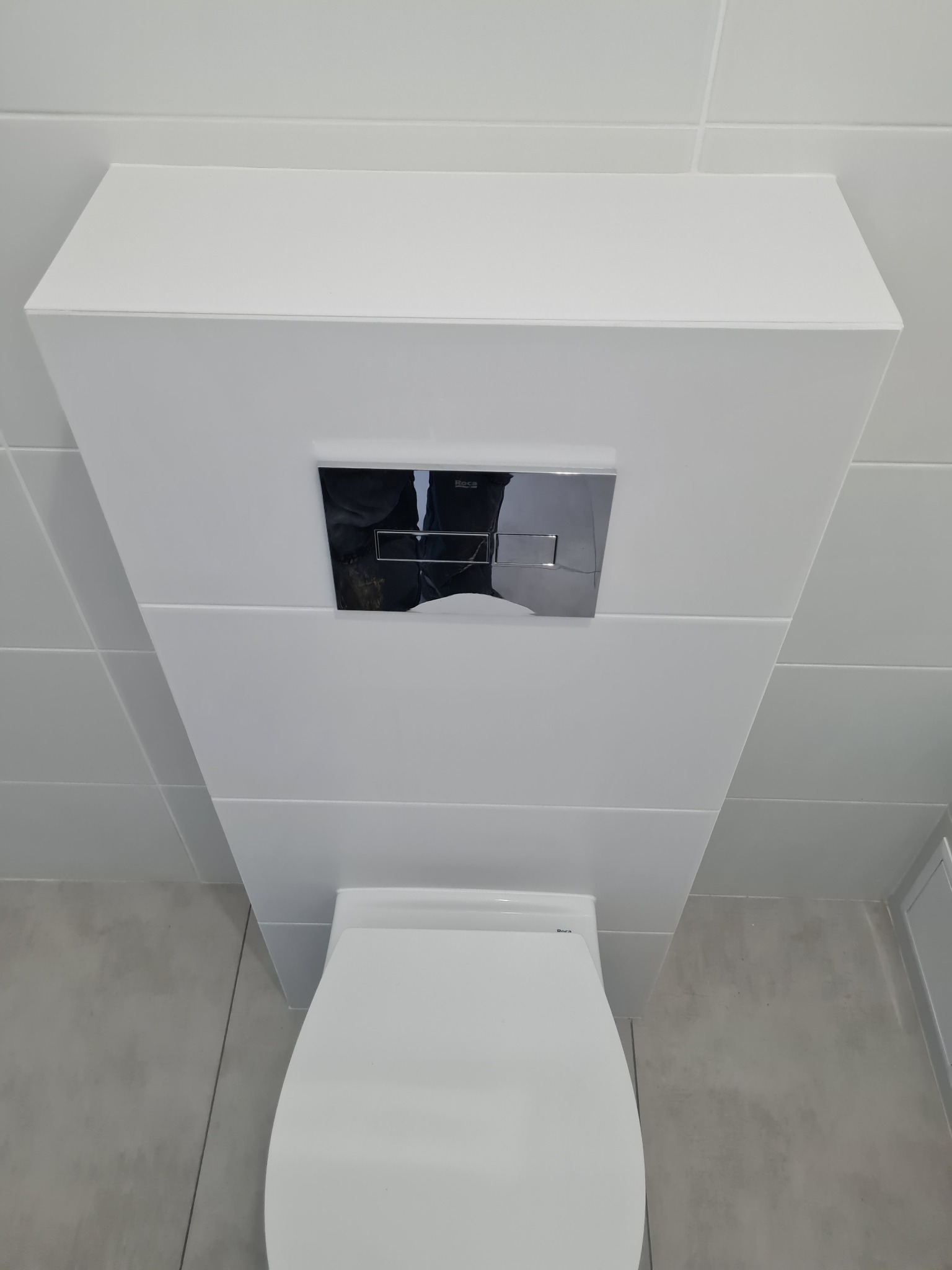 Podwieszana biała toaleta z chromowanym panelem spłukującym, zamontowana na tle jasnych płytek ceramicznych w nowoczesnej łazience.