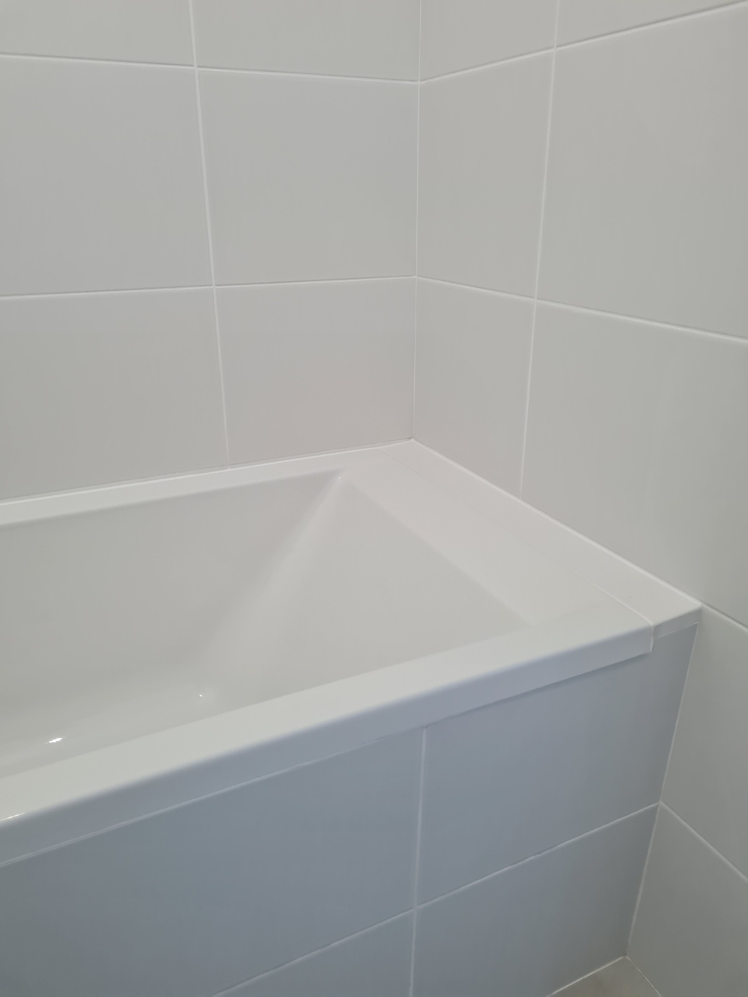 Fragment narożnika łazienki z białą wanną obudowaną płytkami ceramicznymi w jasnym odcieniu szarości, widoczne fugi.