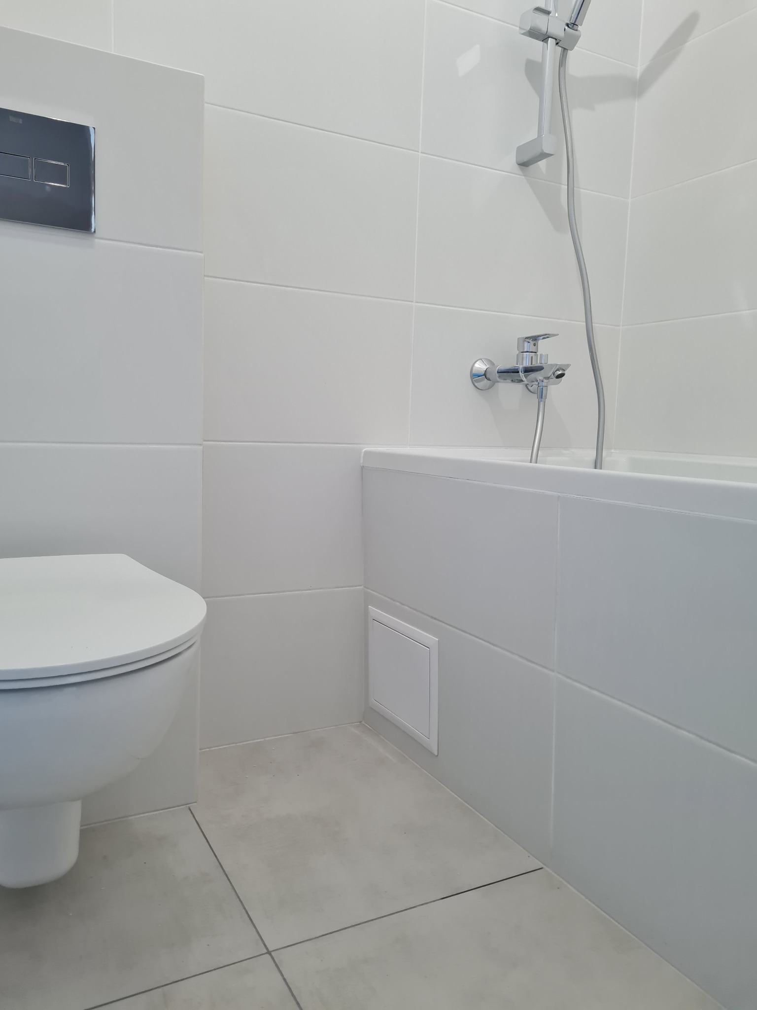 Minimalistyczna, jasna łazienka z wiszącą toaletą, wanną obudowaną płytkami i chromowaną armaturą, wykończona w odcieniach bieli i szarości z matowymi płytkami podłogowymi.