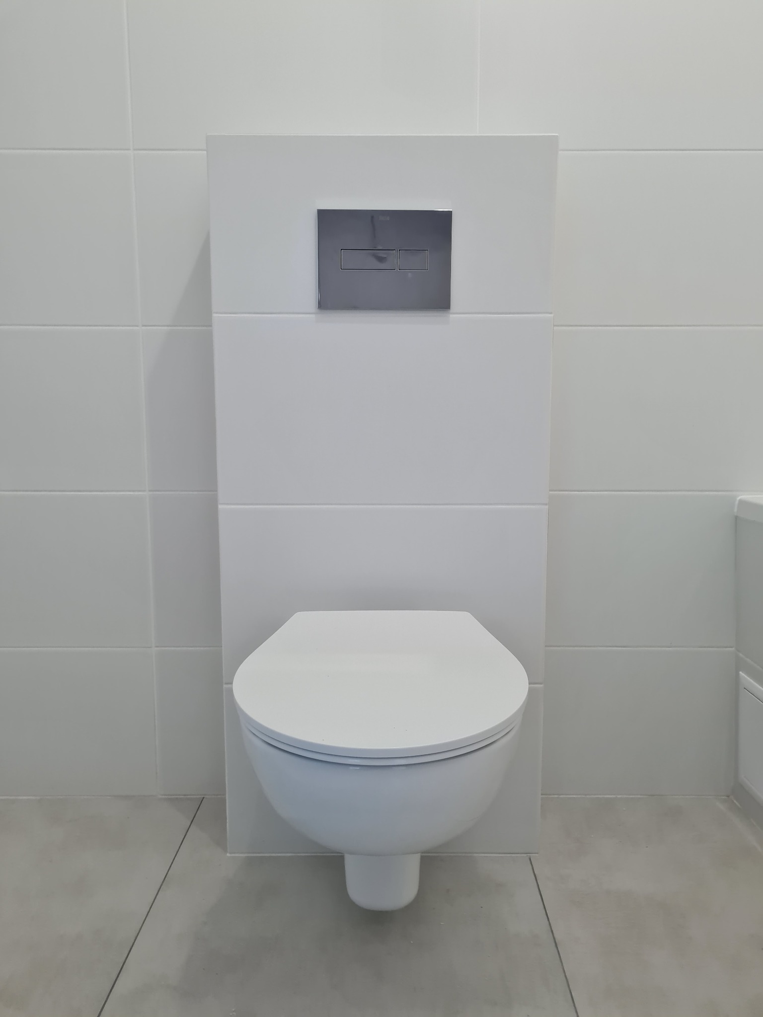 Biała, podwieszana toaleta z chromowanym przyciskiem spłukującym na tle białych płytek w minimalistycznej łazience.