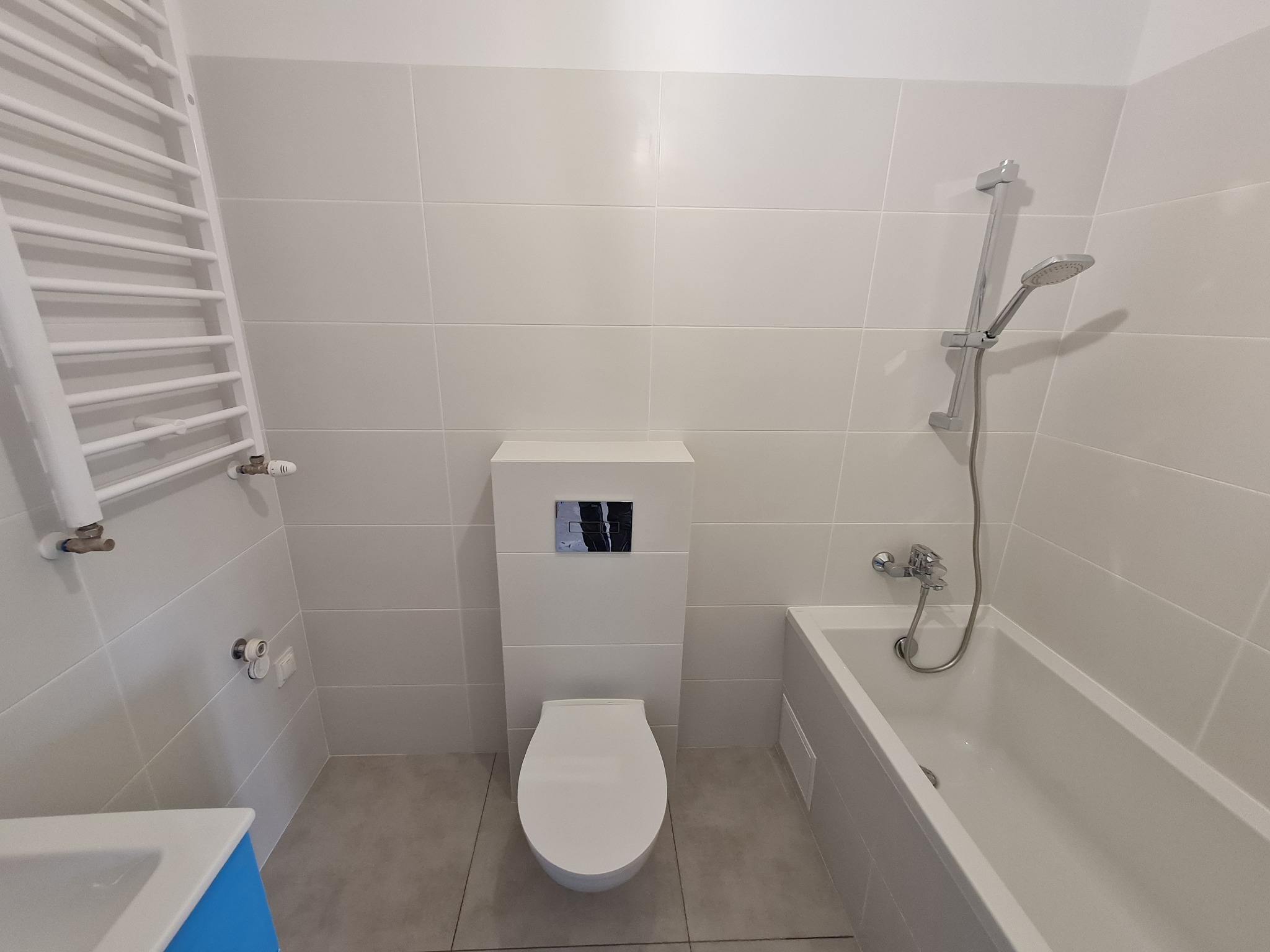 Nowoczesna, jasna łazienka z białą ceramiką, grzejnikiem drabinkowym i wanną ze słuchawką prysznicową, wykończona płytkami w odcieniach szarości.