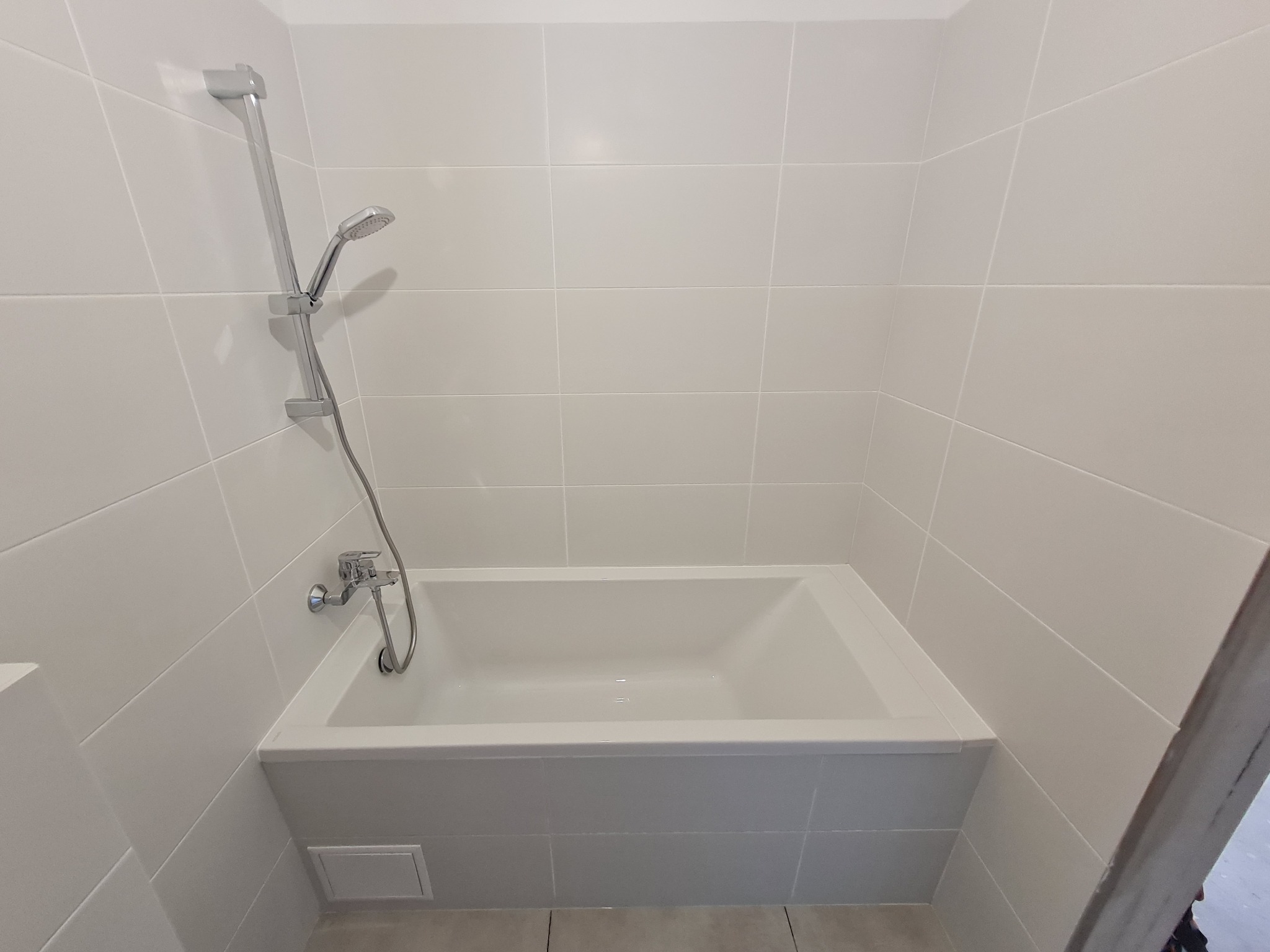 Wanna z obudową wyłożoną białymi płytkami, chromowana bateria i słuchawka prysznicowa na tle ściany wyłożonej tymi samymi płytkami.