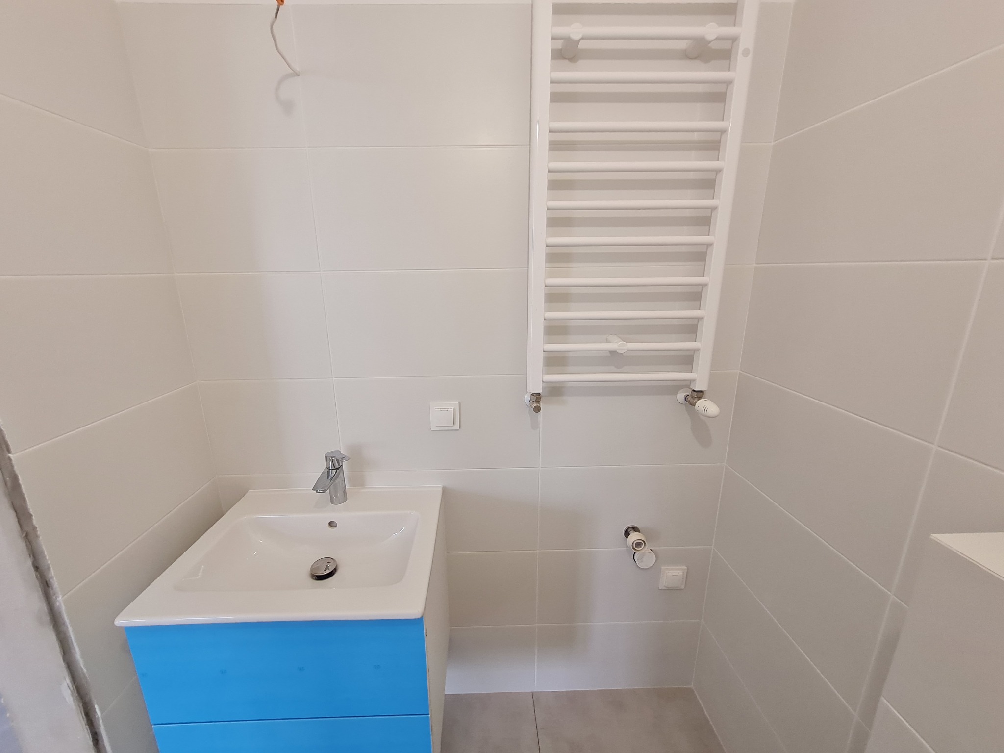 Nowoczesna łazienka z umywalką na niebieskiej szafce, chromowaną baterią i białym grzejnikiem drabinkowym na tle jasnych, kwadratowych płytek.