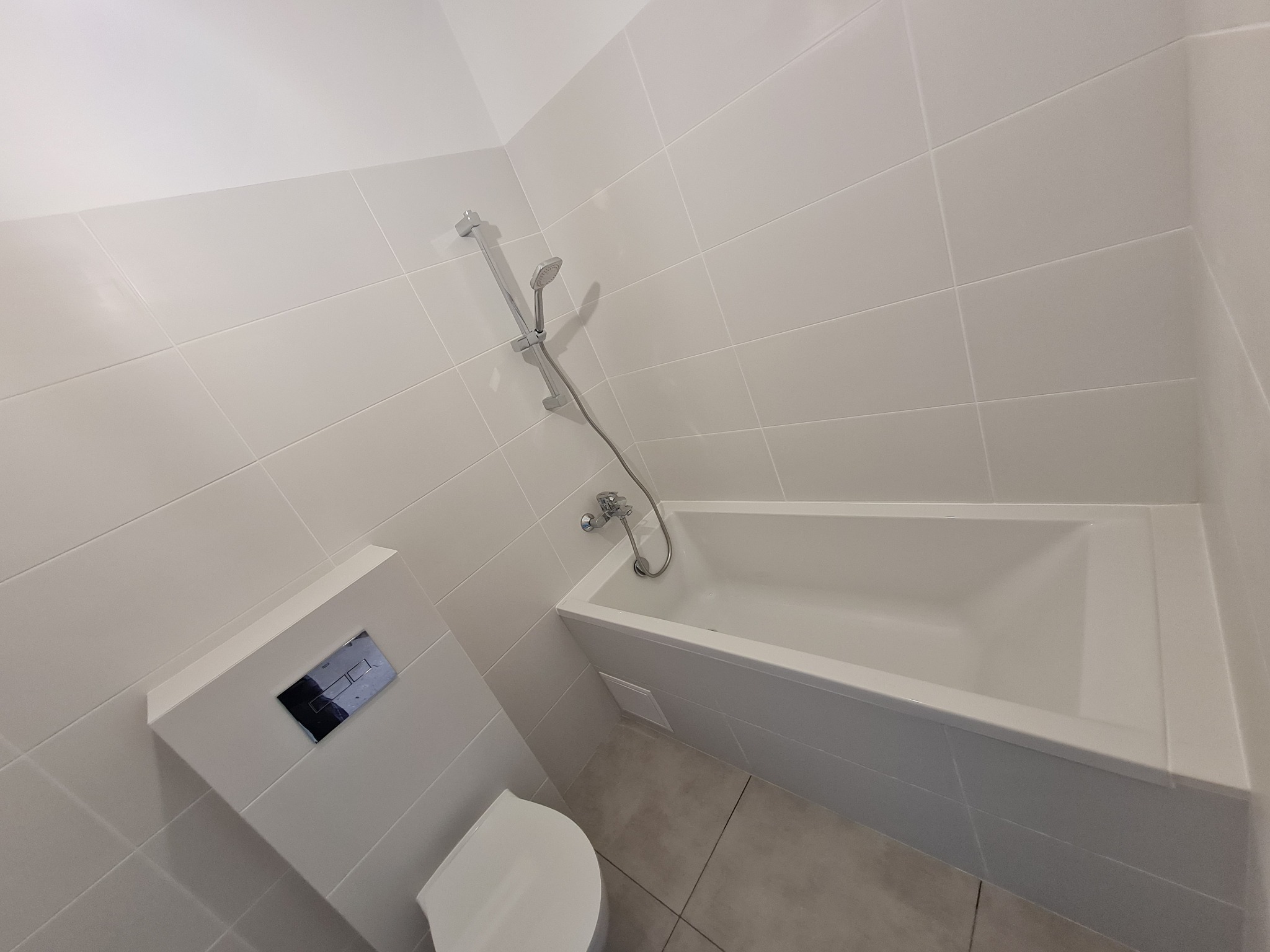 Wykończona łazienka w jasnych płytkach z białą wanną zabudowaną i podwieszanym WC z chromowanym przyciskiem spłukiwania oraz baterią prysznicową.