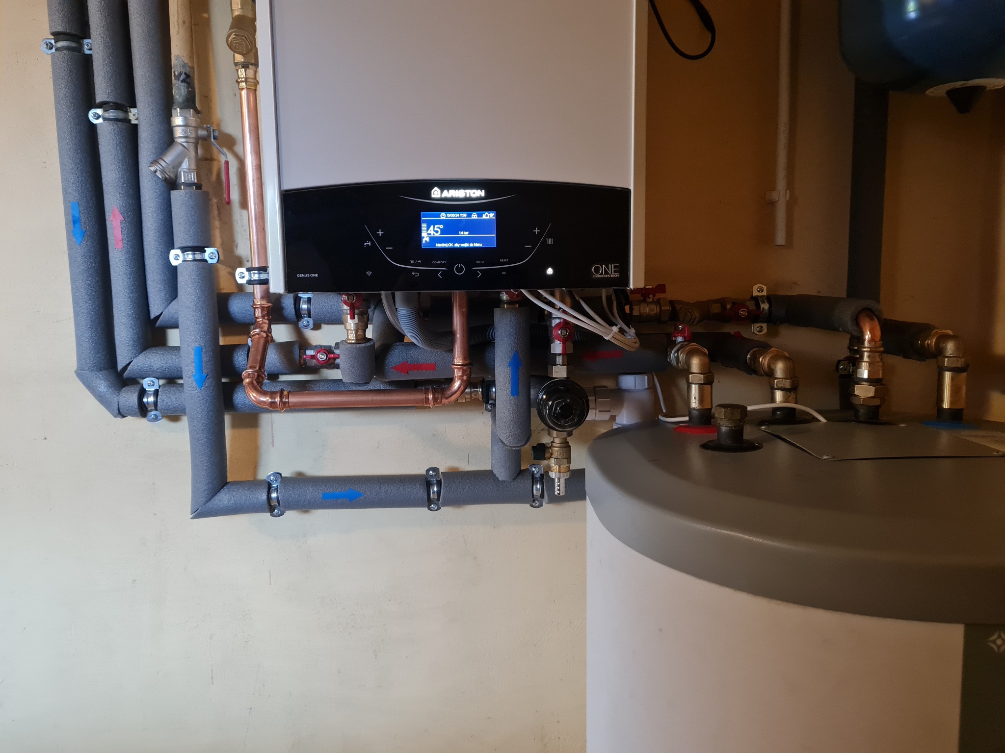 Nowoczesny piec gazowy Ariston z wyświetlaczem, podłączony do instalacji grzewczej z zaizolowanymi rurami oznaczonymi strzałkami kierunku przepływu, obok zasobnik ciepłej wody.
