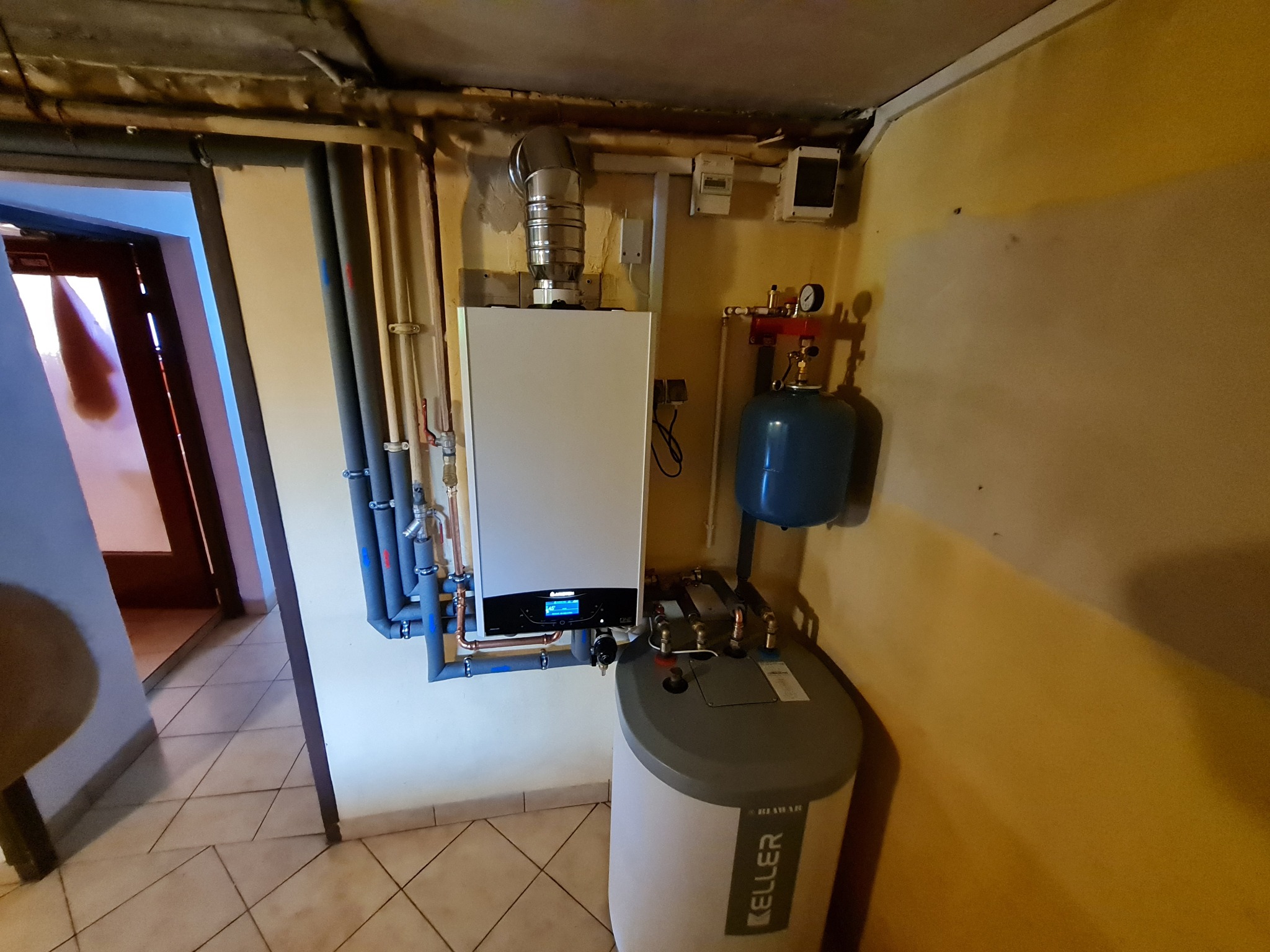 Nowoczesny piec gazowy kondensacyjny z zasobnikiem ciepłej wody, rurami instalacyjnymi w izolacji, zamontowany na żółtej ścianie w piwnicy z płytkami na podłodze.