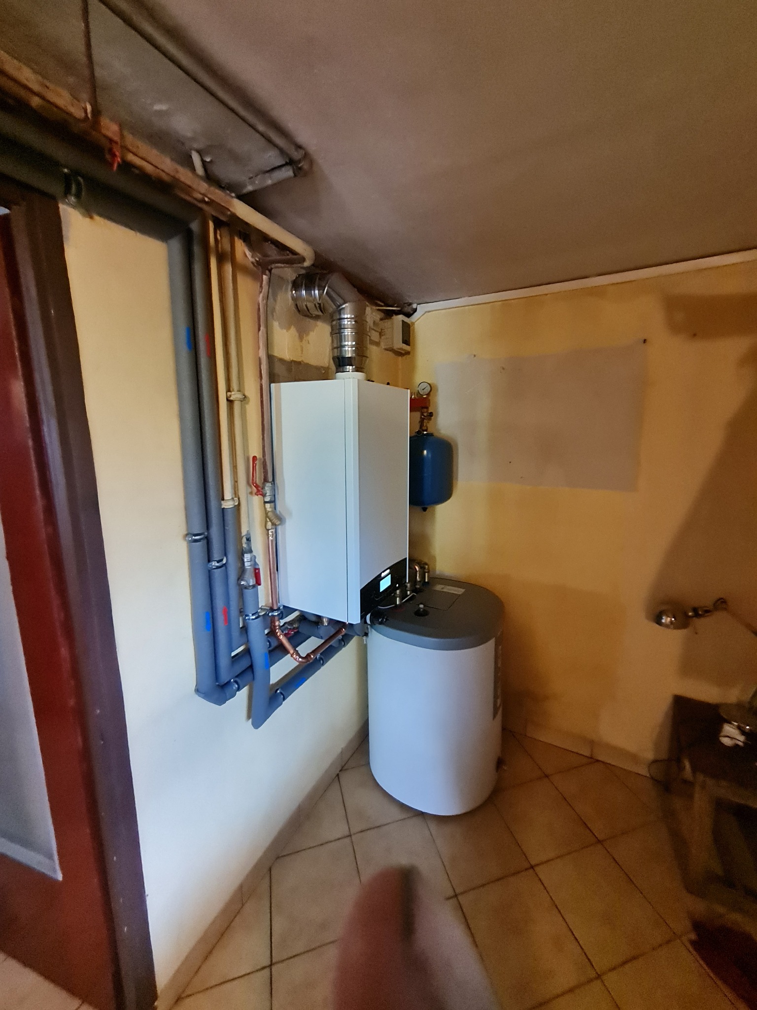 Nowoczesny piec gazowy kondensacyjny z zasobnikiem C.W.U., zamontowany na ścianie w pomieszczeniu technicznym, z widocznymi rurami instalacji grzewczej w izolacji.