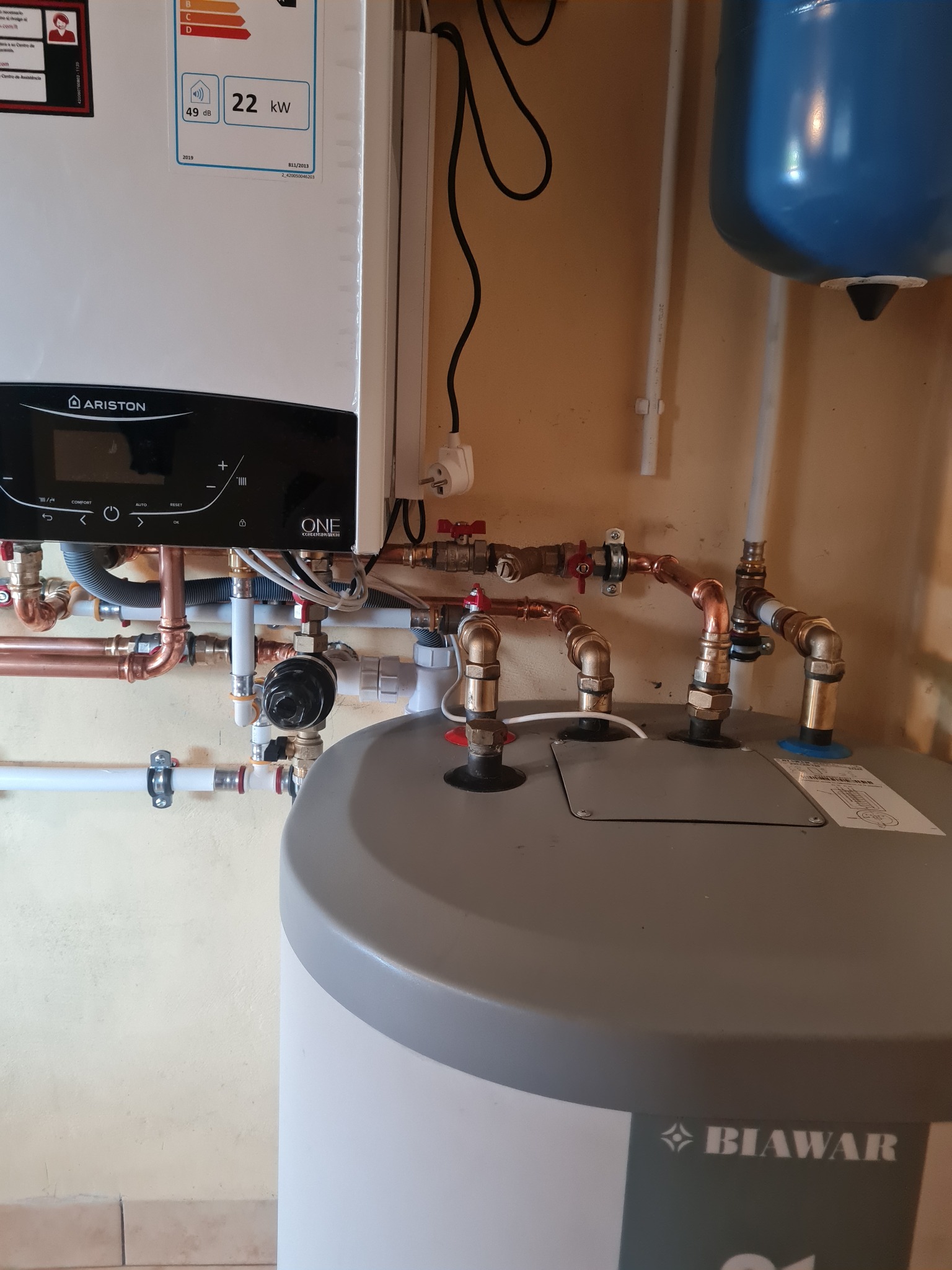 Ariston ONE kondensacyjny piec gazowy z podłączonym zasobnikiem Biawar, widoczne miedziane rury instalacji i elementy sterujące, zamontowane na beżowej ścianie.