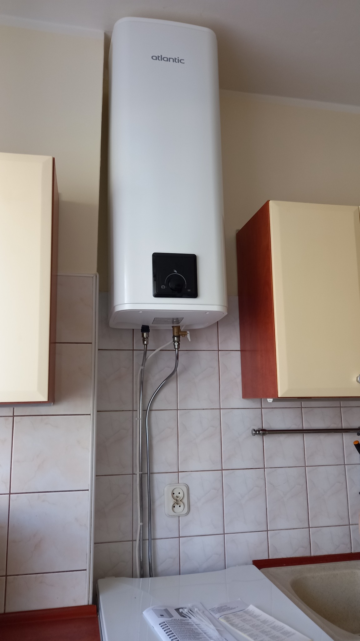 Biały, wiszący elektryczny podgrzewacz wody Atlantic, zamontowany na kafelkowanej ścianie w kuchni, widoczne podłączenia hydrauliczne i szafki kuchenne po bokach.