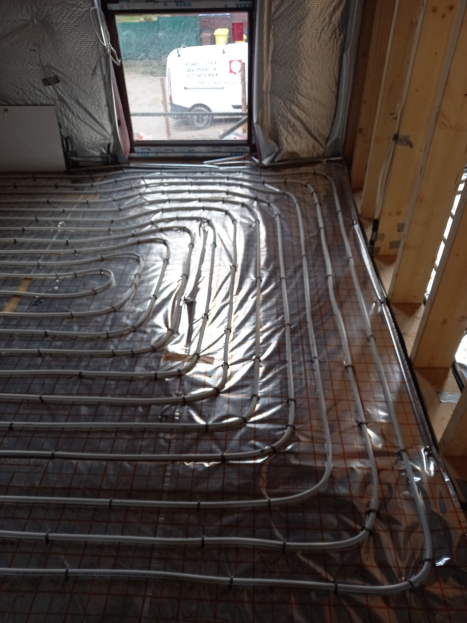 Montaż podłogowego ogrzewania wodnego w nowym budynku, rury rozprowadzone na folii izolacyjnej z widoczną siatką, okno z widokiem na samochód firmy.