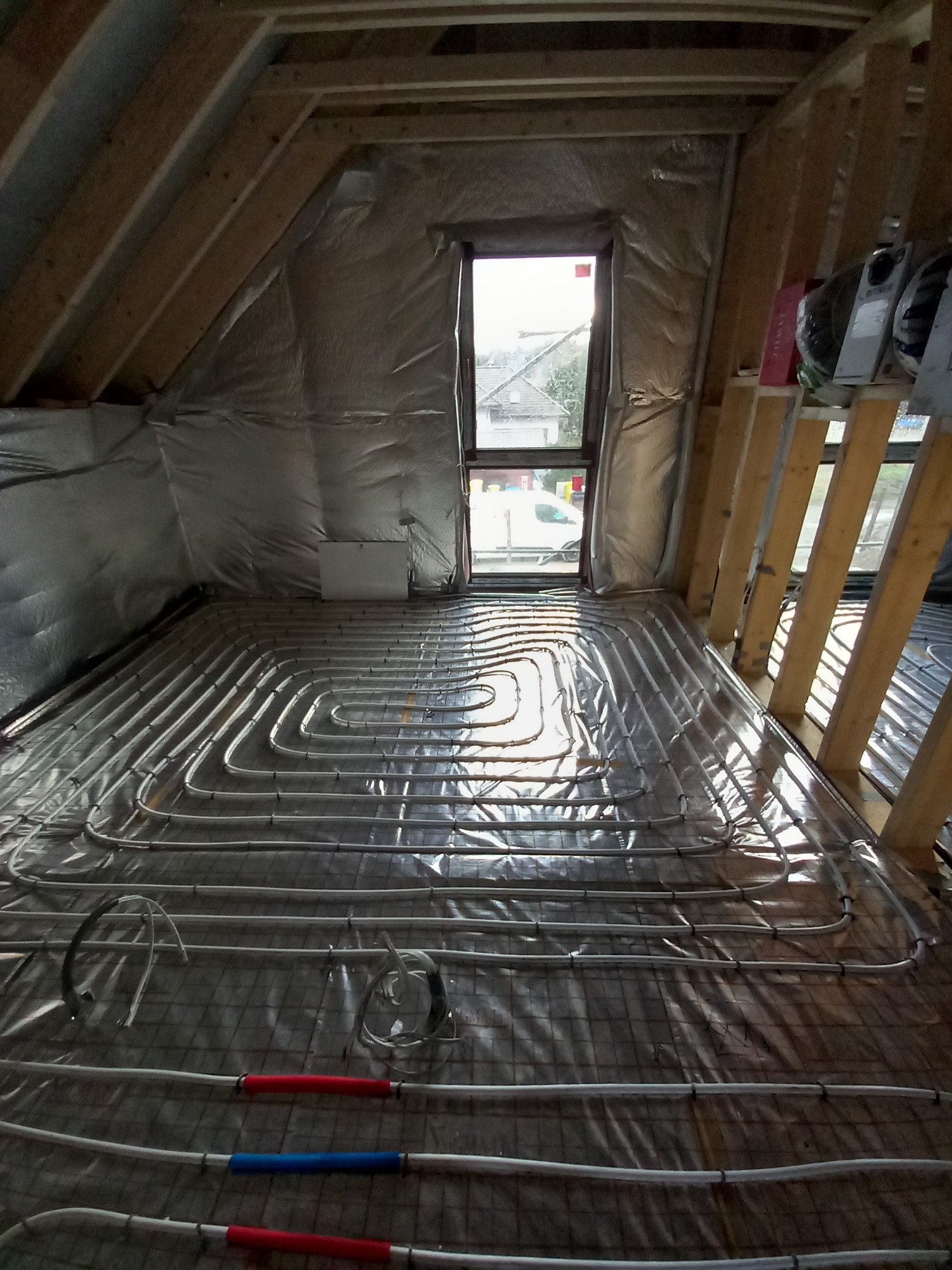 Instalacja ogrzewania podłogowego w nowym domu na poddaszu, widoczne rury w kształcie pętli na folii izolacyjnej, okno w tle.