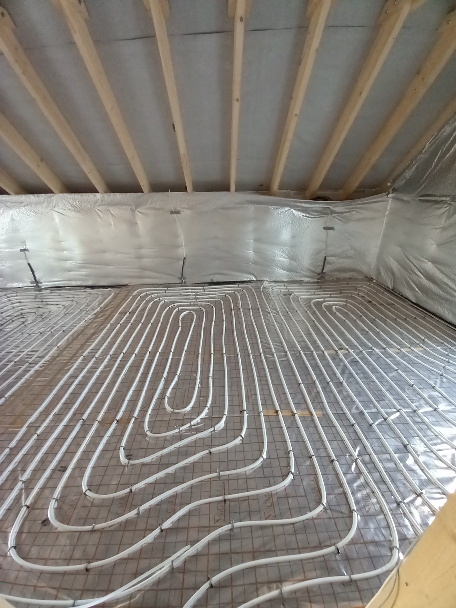 Białe rury ogrzewania podłogowego ułożone w pętle na siatce zbrojeniowej, widok z góry na tle izolacji ścian i drewnianej konstrukcji stropu.