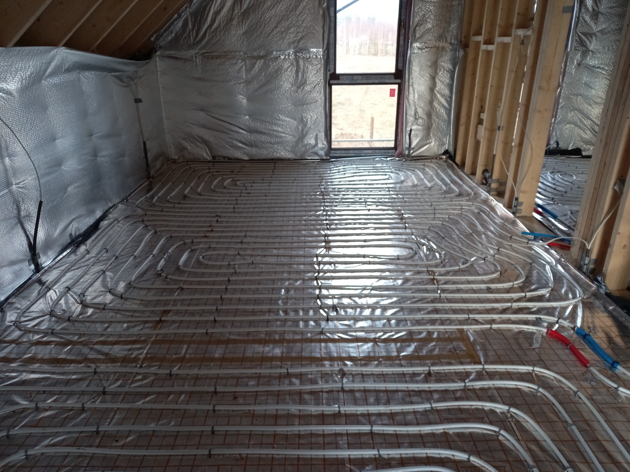 Układanie ogrzewania podłogowego w pomieszczeniu z izolacją termiczną ścian, widoczne białe rurki w pętli oraz drewniana konstrukcja szkieletowa.