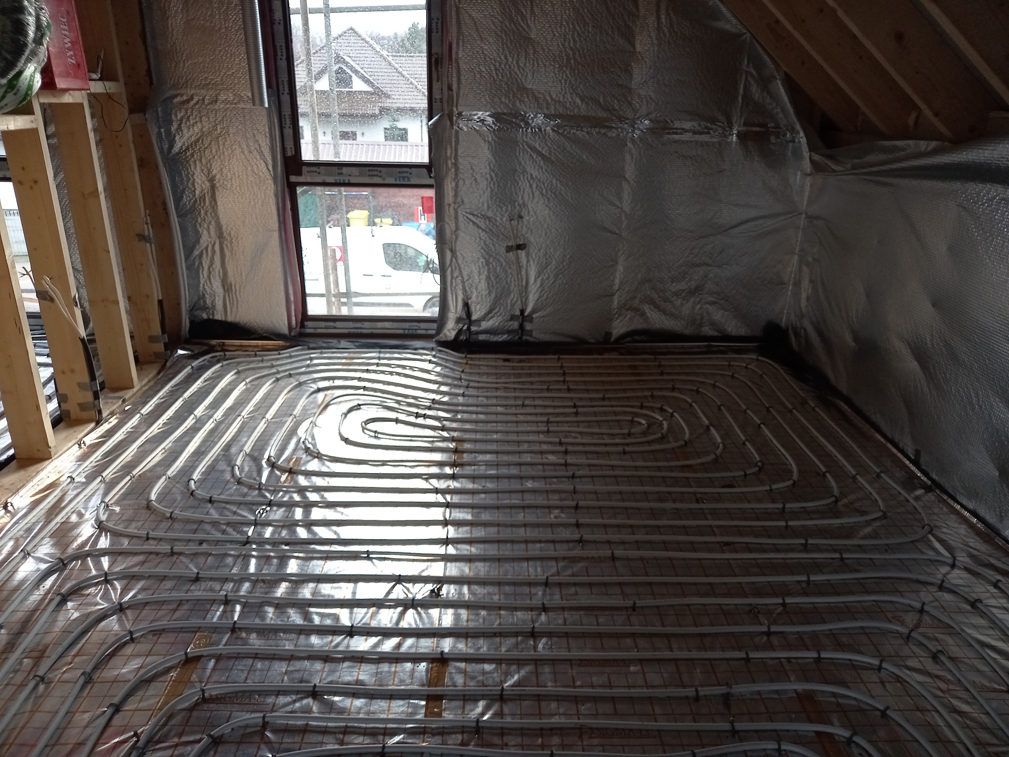 Instalacja ogrzewania podłogowego z rur na siatce, widok z góry, w tle okno z widokiem na dom i samochód dostawczy, ściany ocieplone folią izolacyjną.