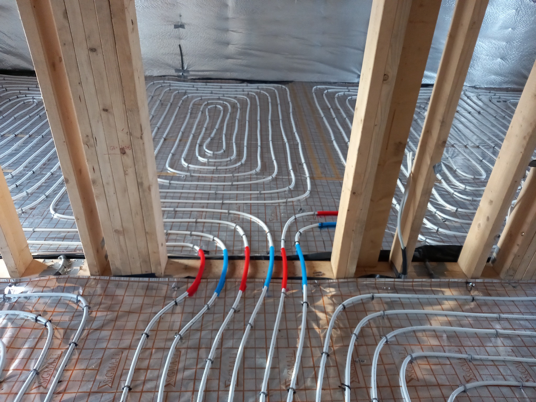 Układanie białych rur ogrzewania podłogowego na folii z siatką, pomiędzy drewnianymi belkami konstrukcyjnymi poddasza, z elementami oznaczonymi na czerwono i niebiesko.