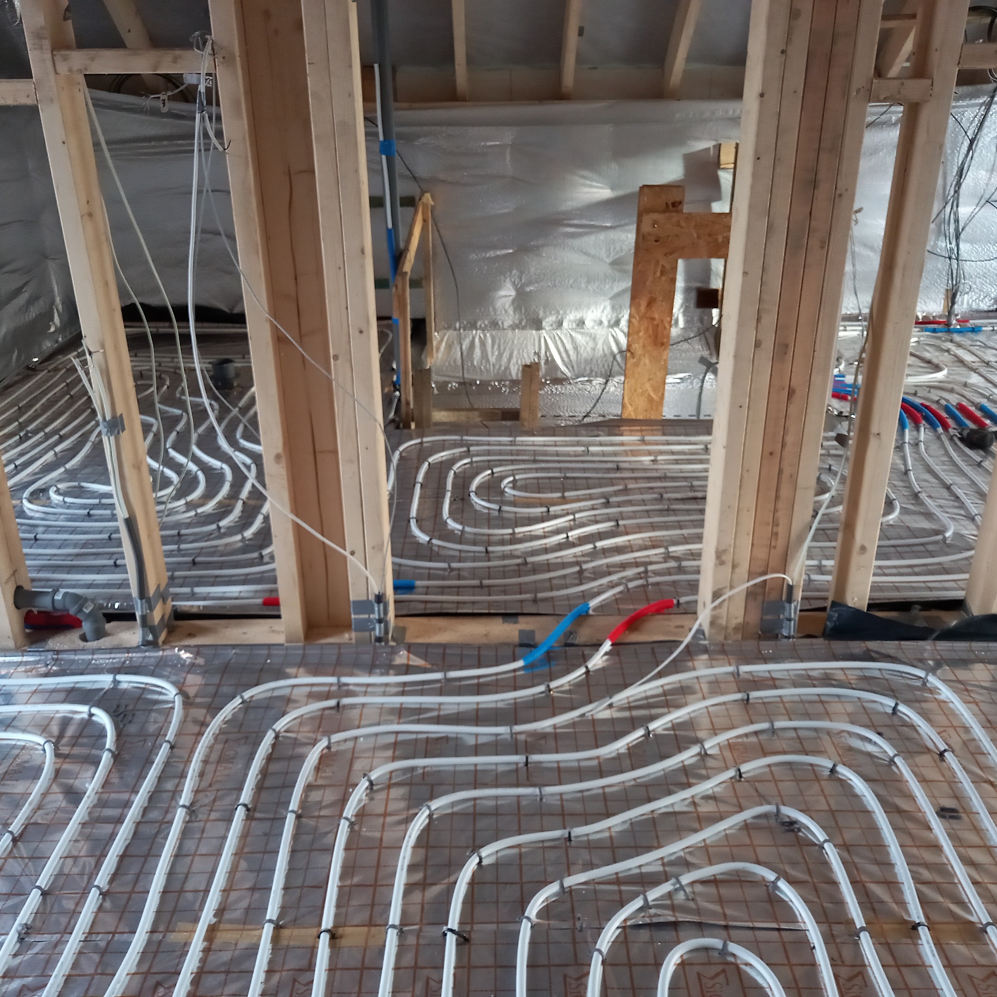 Układanie instalacji ogrzewania podłogowego w Gdańsku, widoczne rury grzewcze zamocowane do siatki na izolacji, drewniane elementy konstrukcyjne budynku w tle.