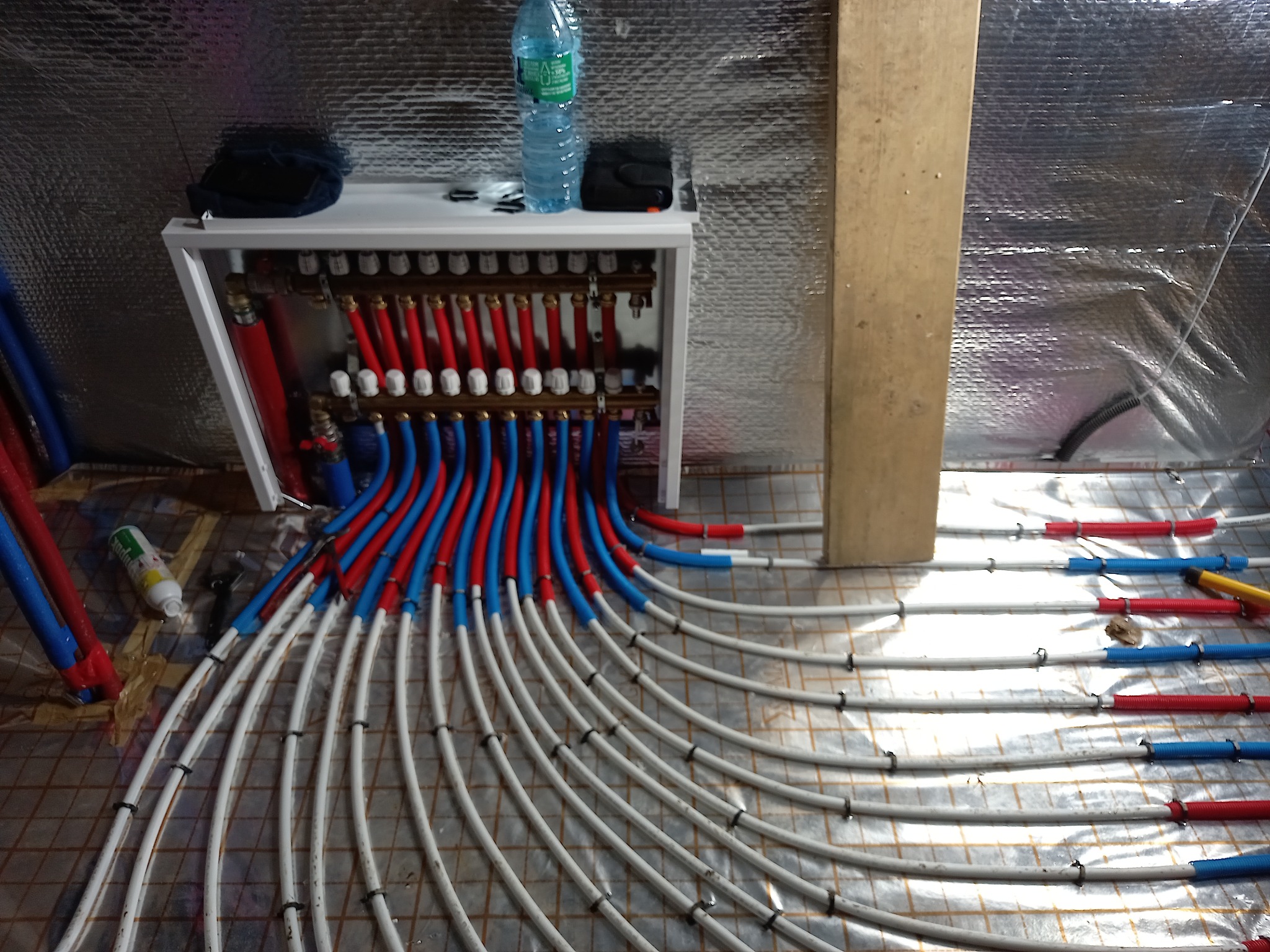 Rozdzielacz ogrzewania podłogowego z podłączonymi rurami, widoczne narzędzia i butelka wody na rozdzielaczu, instalacja w trakcie montażu.