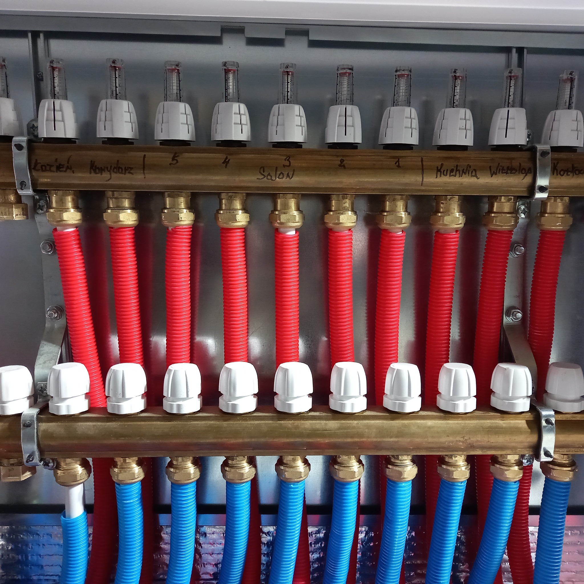 Rozdzielacz ogrzewania podłogowego z czerwonymi i niebieskimi rurami, opisany nazwami pomieszczeń, z widocznymi przepływomierzami.