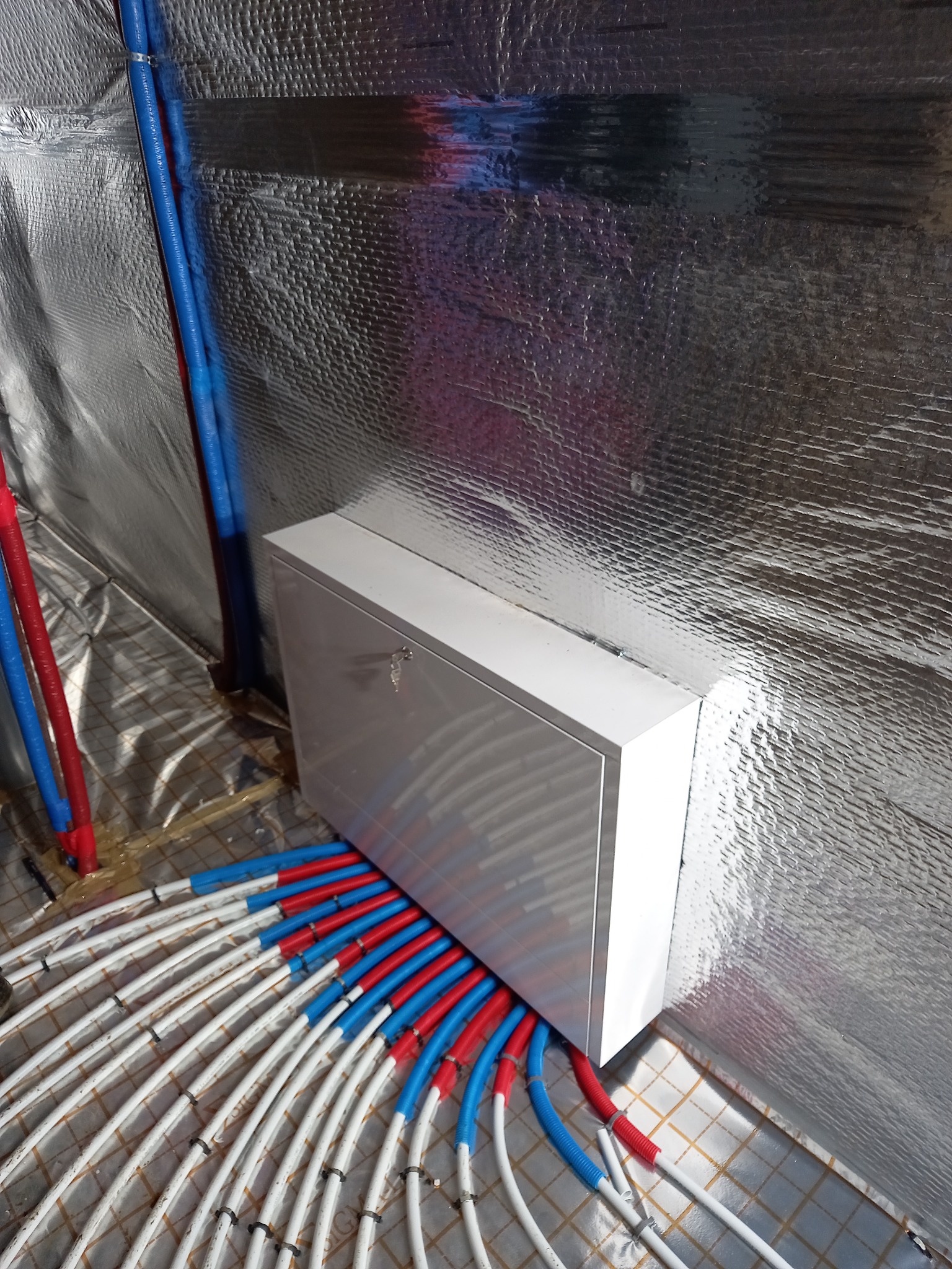 Rozdzielacz ogrzewania podłogowego w białej szafce, z licznymi rurami w kolorach czerwonym i niebieskim, ułożonymi na siatce izolacyjnej, na tle ściany pokrytej folią aluminiową.