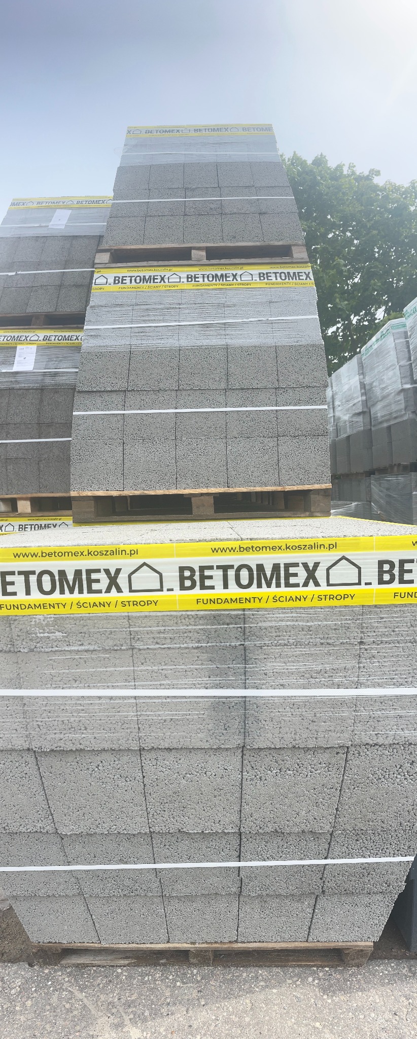 Palety z szarymi bloczkami z betonu komórkowego Betomex, owinięte folią i taśmą z logo producenta, składowane na placu budowy.