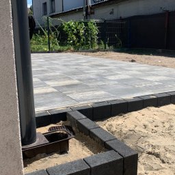 Valerii Radchuk - Ułożona szara kostka brukowa z czarnym obrzeżem w trakcie budowy, widoczny odpływ liniowy i piasek, w tle dom z ogrodem.