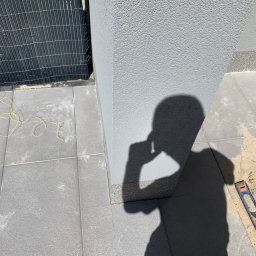 Valerii Radchuk - Szare płyty chodnikowe ułożone przy ścianie budynku, widoczny cień osoby trzymającej telefon, poziomica i piasek w spoinach, żółty sznurek na płytach.