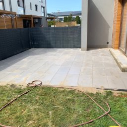 Valerii Radchuk - Nowoczesny taras z jasnych, kwadratowych płyt, częściowo obsypanych piaskiem, z widocznym wężem ogrodowym na trawie i fragmentem drewnianej elewacji budynku.