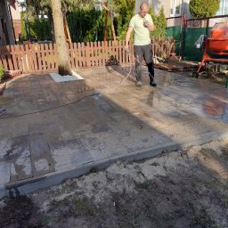 Valerii Radchuk - Mężczyzna czyści kostkę brukową szarą wodą pod ciśnieniem, obok stoi pomarańczowa betoniarka, w tle drewniany płot i drzewa.