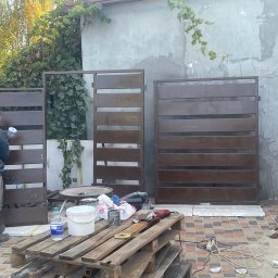 Valerii Radchuk - Malowanie brązowej, metalowej bramy ogrodzeniowej pistoletem natryskowym na zewnątrz, widoczny paleta z farbami, szlifierka kątowa i niebieski parasol w tle.