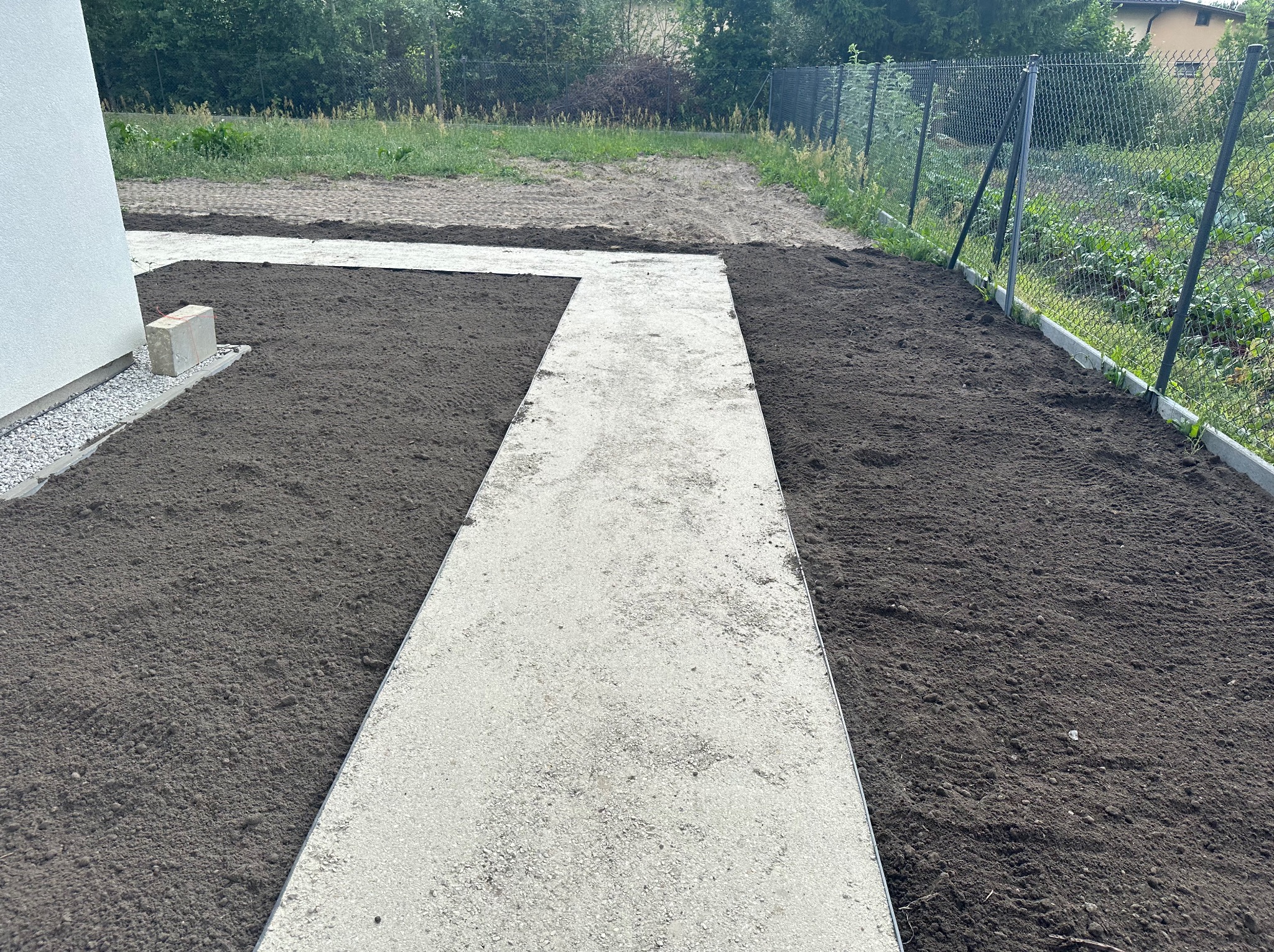 Świeżo ułożony, jasnoszary chodnik z płyt betonowych prowadzący przez wyrównany teren z czarną ziemią, widoczne ogrodzenie z siatki i fragment budynku.