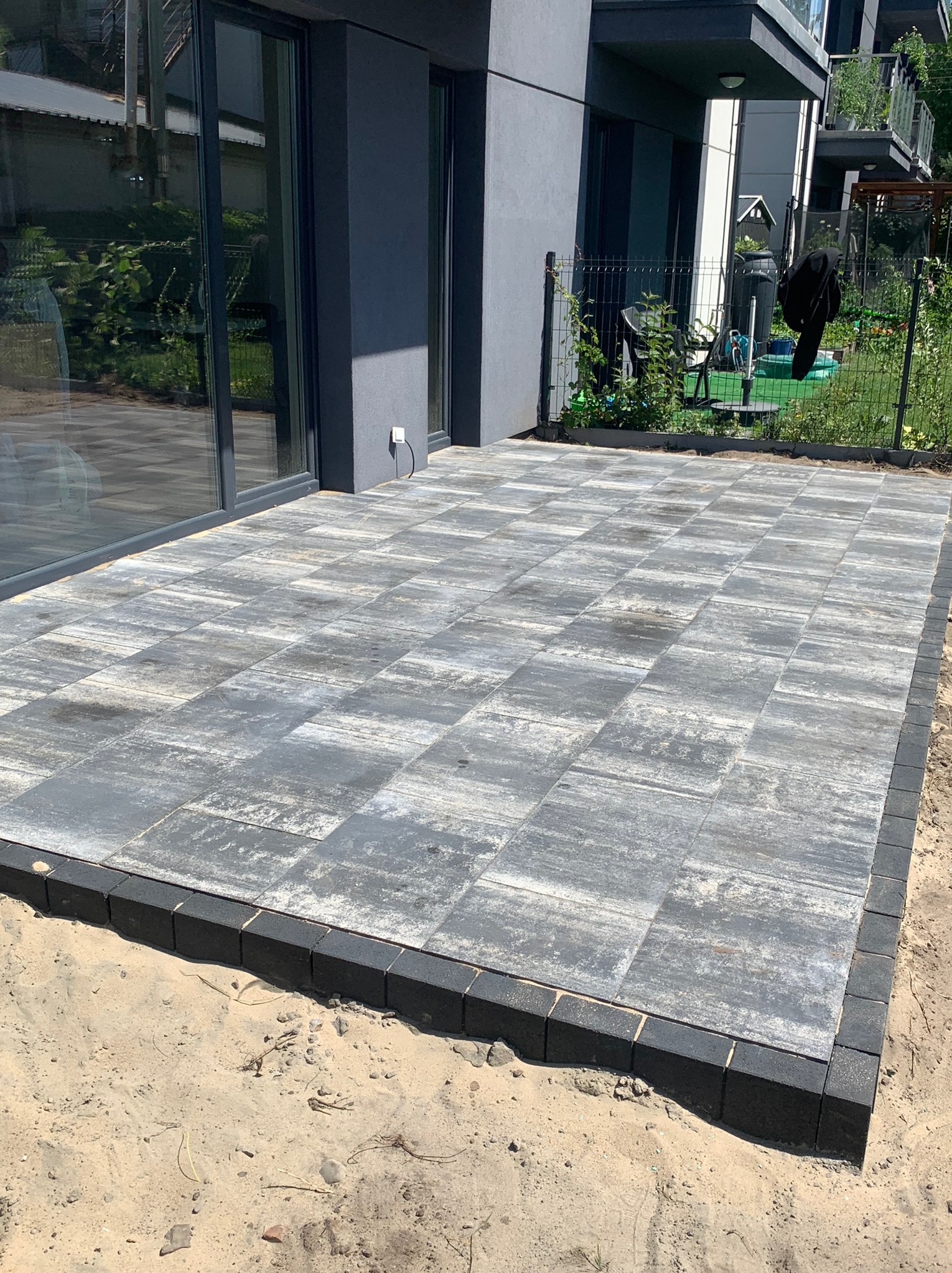 Ułożona szara kostka brukowa na tarasie przed nowoczesnym budynkiem mieszkalnym, obrzeża z ciemnej kostki, widoczny piasek wokół, w tle ogród za ogrodzeniem.