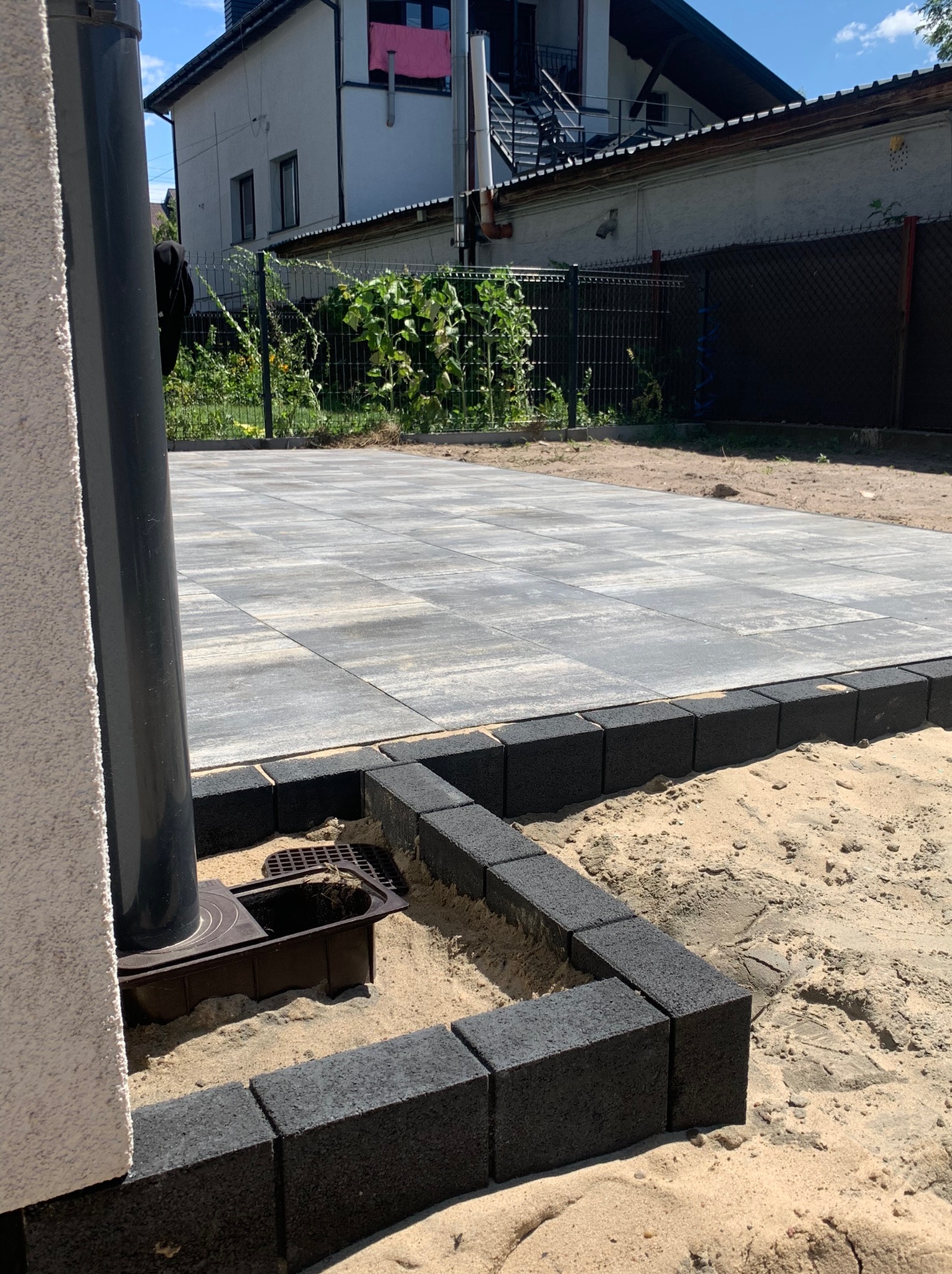 Ułożona szara kostka brukowa z czarnym obrzeżem w trakcie budowy, widoczny odpływ liniowy i piasek, w tle dom z ogrodem.