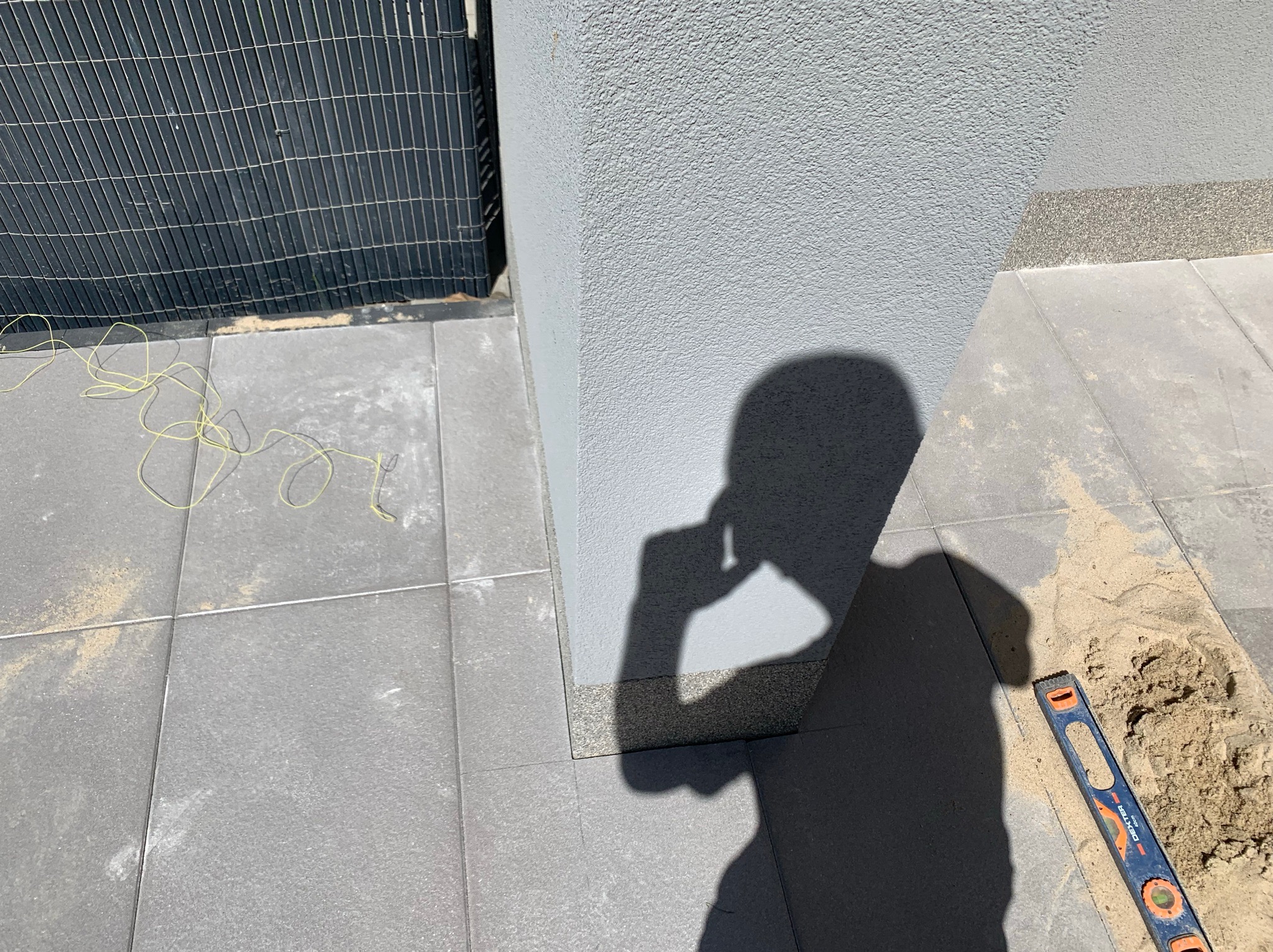 Szare płyty chodnikowe ułożone przy ścianie budynku, widoczny cień osoby trzymającej telefon, poziomica i piasek w spoinach, żółty sznurek na płytach.