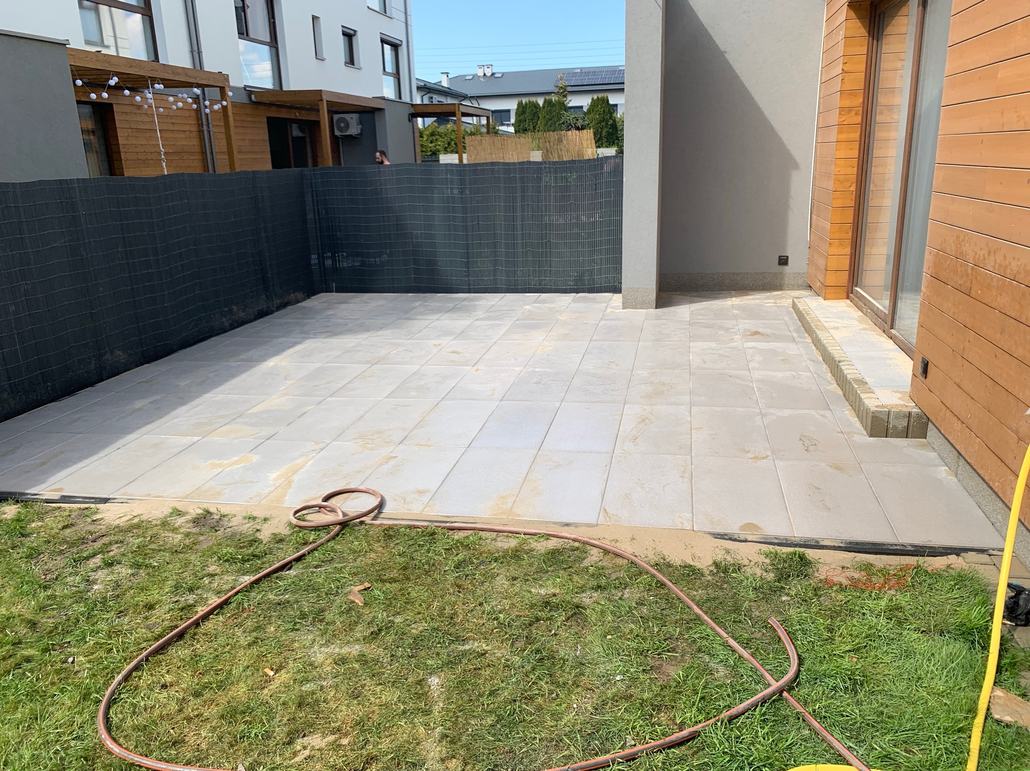 Nowoczesny taras z jasnych, kwadratowych płyt, częściowo obsypanych piaskiem, z widocznym wężem ogrodowym na trawie i fragmentem drewnianej elewacji budynku.