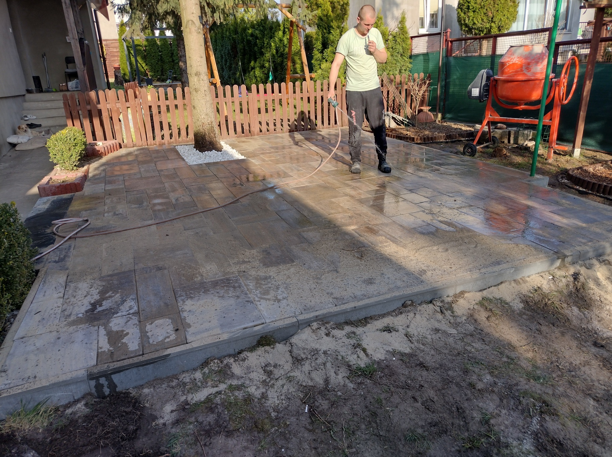 Mężczyzna czyści kostkę brukową szarą wodą pod ciśnieniem, obok stoi pomarańczowa betoniarka, w tle drewniany płot i drzewa.