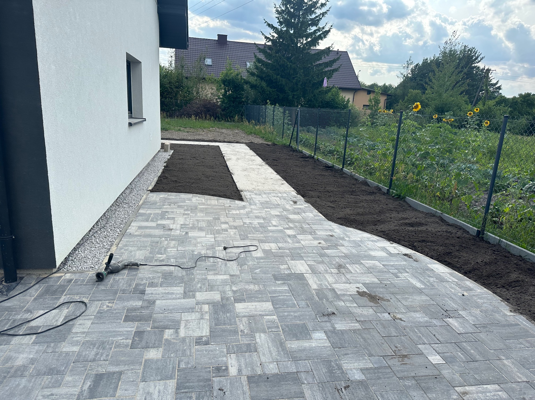 Ułożona szara kostka brukowa na podwórku nowego domu, z przygotowanymi rabatami ziemnymi wzdłuż ogrodzenia i budynku, oraz szlifierką kątową leżącą na kostce.