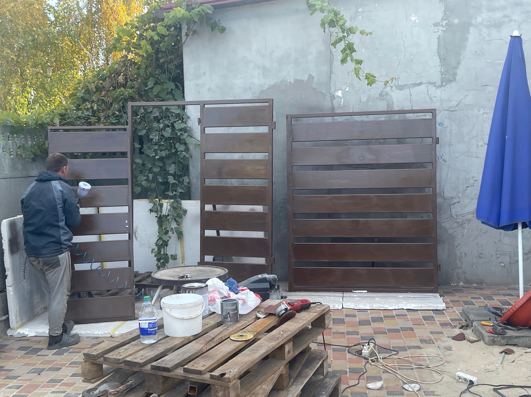 Malowanie brązowej, metalowej bramy ogrodzeniowej pistoletem natryskowym na zewnątrz, widoczny paleta z farbami, szlifierka kątowa i niebieski parasol w tle.