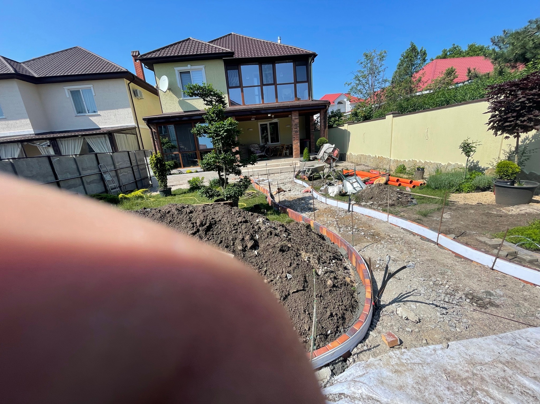 Przygotowanie podłoża pod ścieżkę z obrzeżami z cegły klinkierowej, częściowo zasypane ziemią, widoczne narzędzia i dom w tle, ujęcie z bliska z elementem ciała w kadrze.