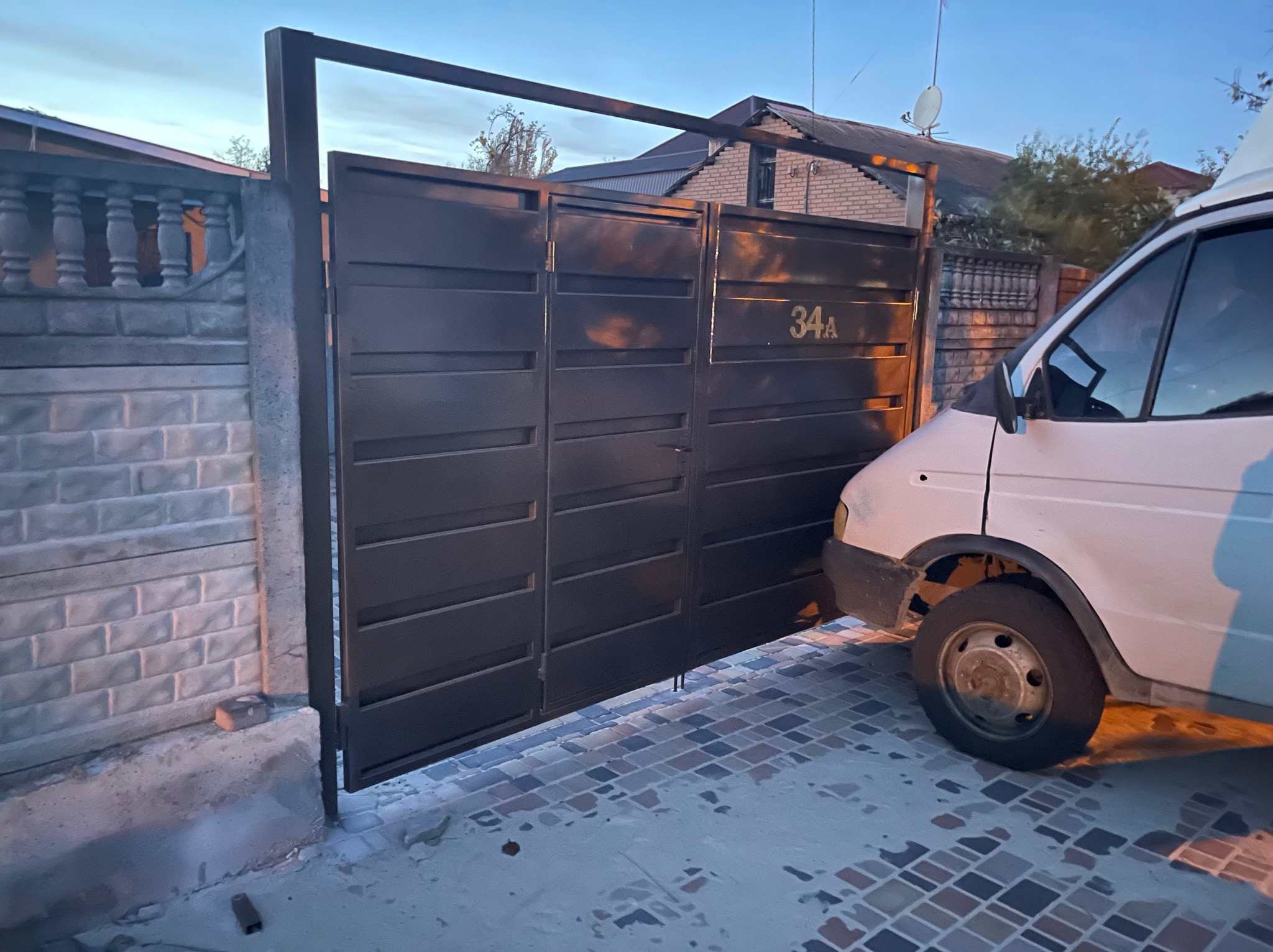 Ciemna, pozioma brama z numerem 34A, zamontowana na betonowych słupkach, widoczna z perspektywy podjazdu wyłożonego kostką w odcieniach szarości i brązu, z zaparkowanym białym samochodem dostawczym...