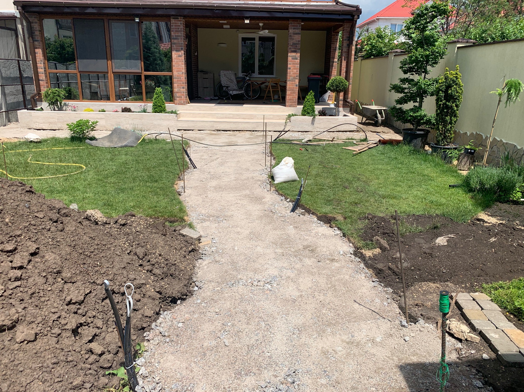 Przygotowanie podłoża pod ścieżkę ogrodową z użyciem tłucznia, widoczne paliki wyznaczające trasę, obok pryzma ziemi i narzędzia, w tle dom z tarasem i zadbany ogród.