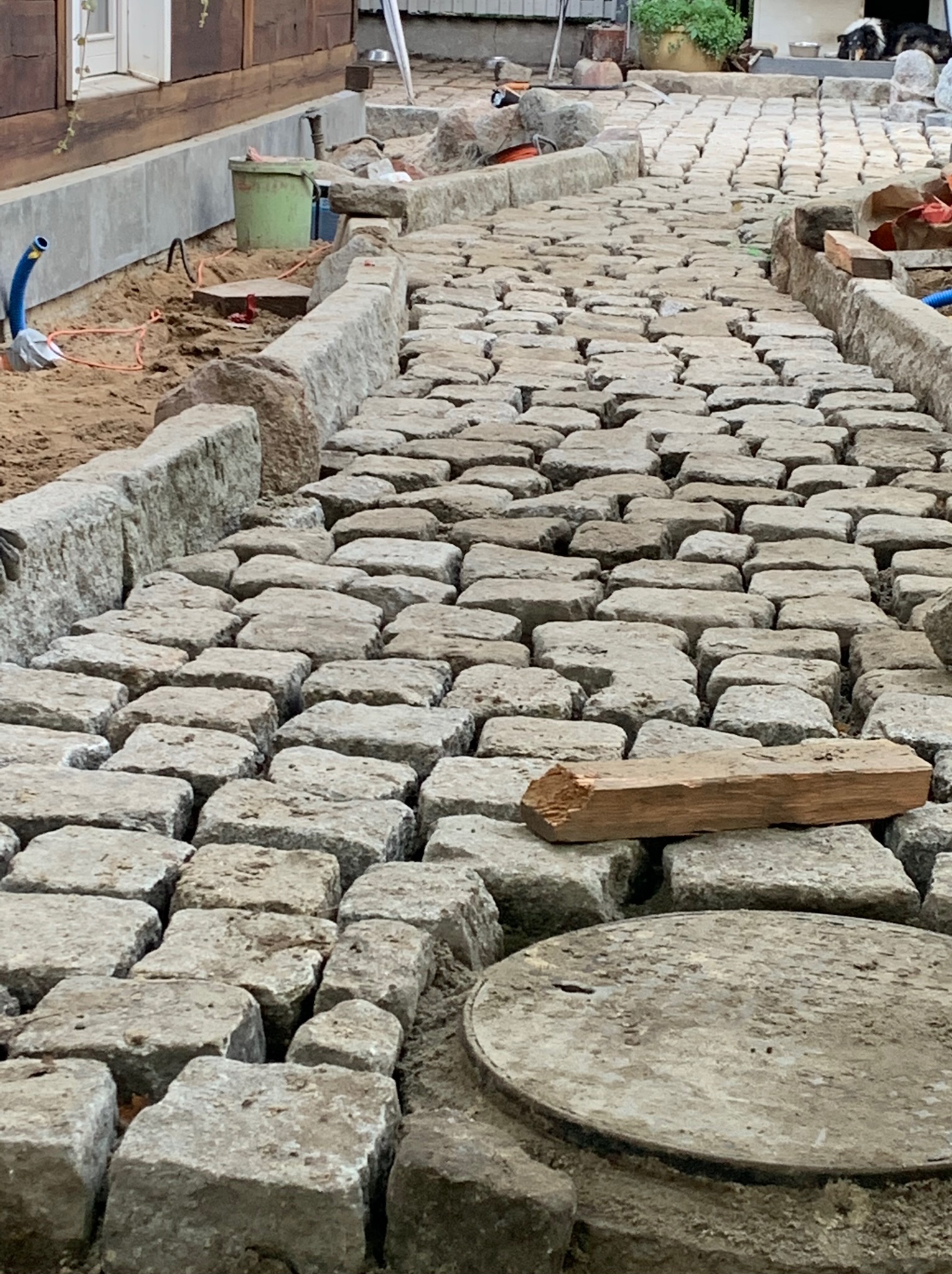 Ścieżka z kostki granitowej w trakcie układania, widoczny fragment drewnianego klocka na powierzchni, kanalizacja pod spodem, obrzeża z kamienia.