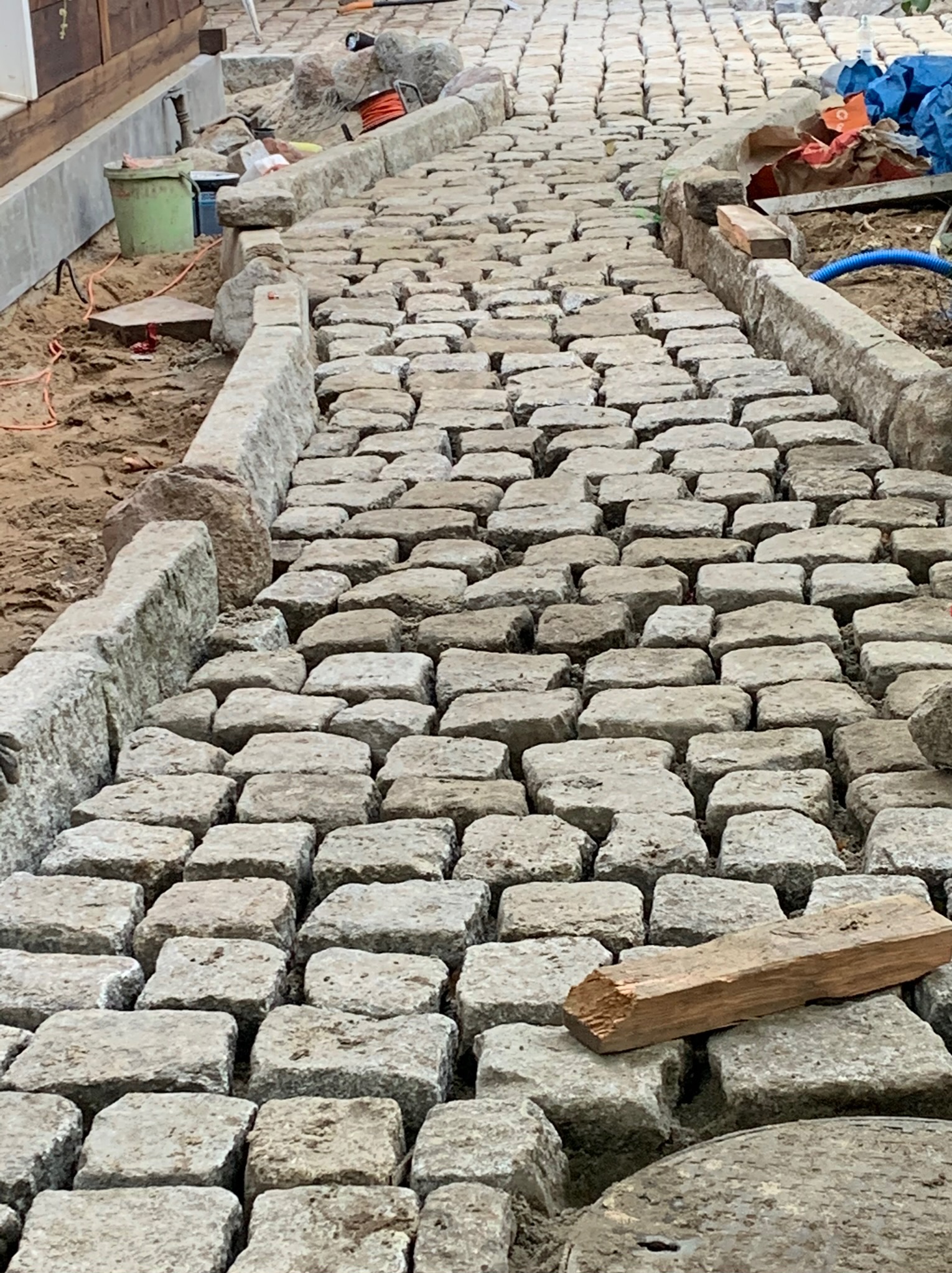 Ścieżka z jasnoszarej kostki granitowej, obramowana betonowymi krawężnikami, w trakcie budowy; w tle widoczne narzędzia i materiały budowlane.