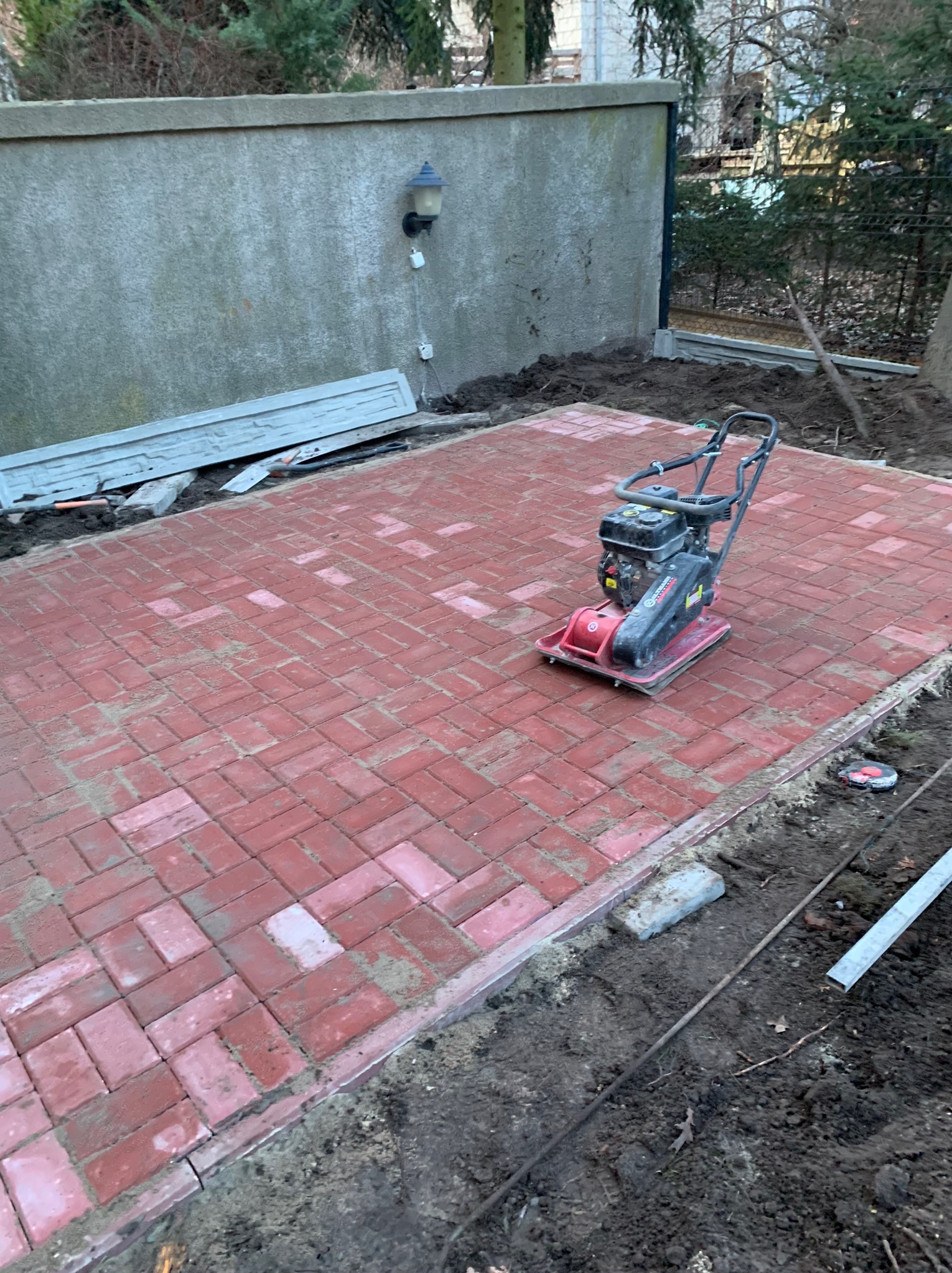 Czerwona kostka brukowa ułożona na podwórku, zagęszczarka spalinowa stoi na powierzchni, obok widoczna ziemia i fragmenty obrzeży, w tle szary mur i drzewa.