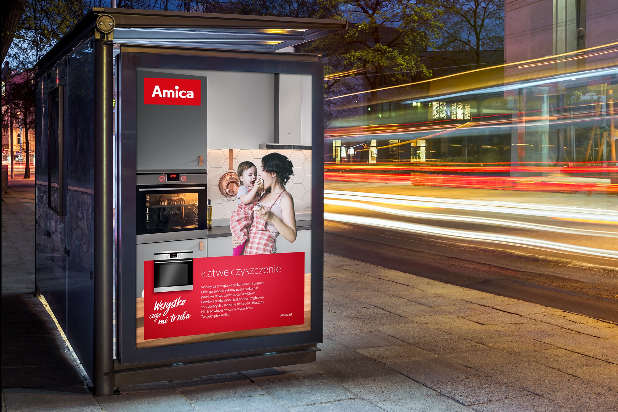 Przystanek autobusowy z podświetlonym plakatem reklamowym piekarników Amica, rozmyte światła przejeżdżających pojazdów w tle.
