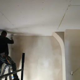Pracownik na drabinie szpachluje sufit z płyt gipsowo-kartonowych w pomieszczeniu z łukowym przejściem, widoczne łączenia płyt i nierówności na ścianie.