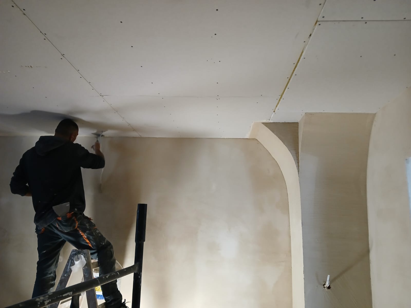 Pracownik na drabinie szpachluje sufit z płyt gipsowo-kartonowych w pomieszczeniu z łukowym przejściem, widoczne łączenia płyt i nierówności na ścianie.